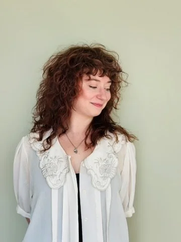 A woman with curly red hair, wearing a white blouse with intricate embroidery and a necklace, standing against a light green wall with her eyes closed and a slight smile.