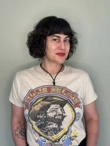 Woman with dark curly hair wearing a graphic t-shirt with a portrait and a necklace, standing against a pale green wall.