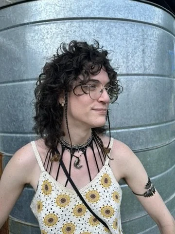 Young person with curly dark hair and glasses standing outdoors in front of a galvanized metal water tank, wearing a sunflower-patterned dress and layered necklaces.