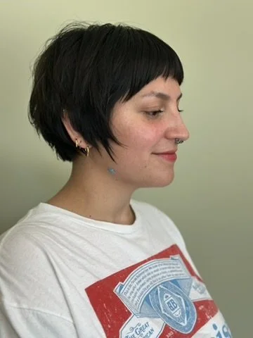 Profile of a young woman with short black hair, wearing a white t-shirt with a colorful graphic design, standing against a light green background.