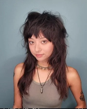 Young woman with messy, voluminous, dark hair, wearing a gray tank top, layered necklaces, and a tattoo on her left arm, standing against a plain blue background.