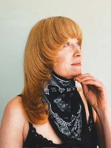 Profile of a woman with red hair wearing a black bandana and a black top, touching her chin.