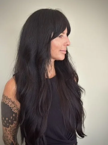 Profile of a woman with long black hair, wearing a sleeveless black top, against a plain light background.