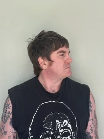 Side profile of a young man with tattoos on his arms, wearing a black sleeveless shirt with a white graphic design.