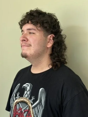 Side profile of a young man with curly dark hair, facial hair, wearing a black T-shirt with a graphic design, standing against a plain light-colored background.