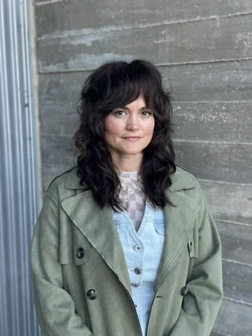 A woman with dark wavy hair in a green jacket standing outdoors against a wooden wall.
