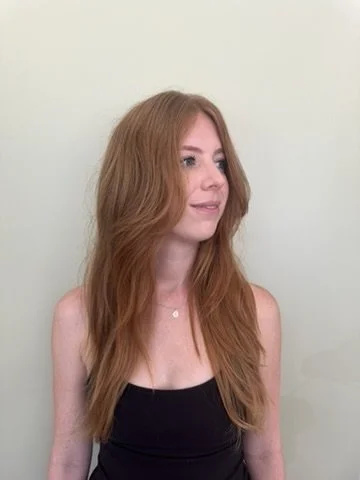 Young woman with long, wavy red hair wearing a black sleeveless top, standing against a light-colored wall