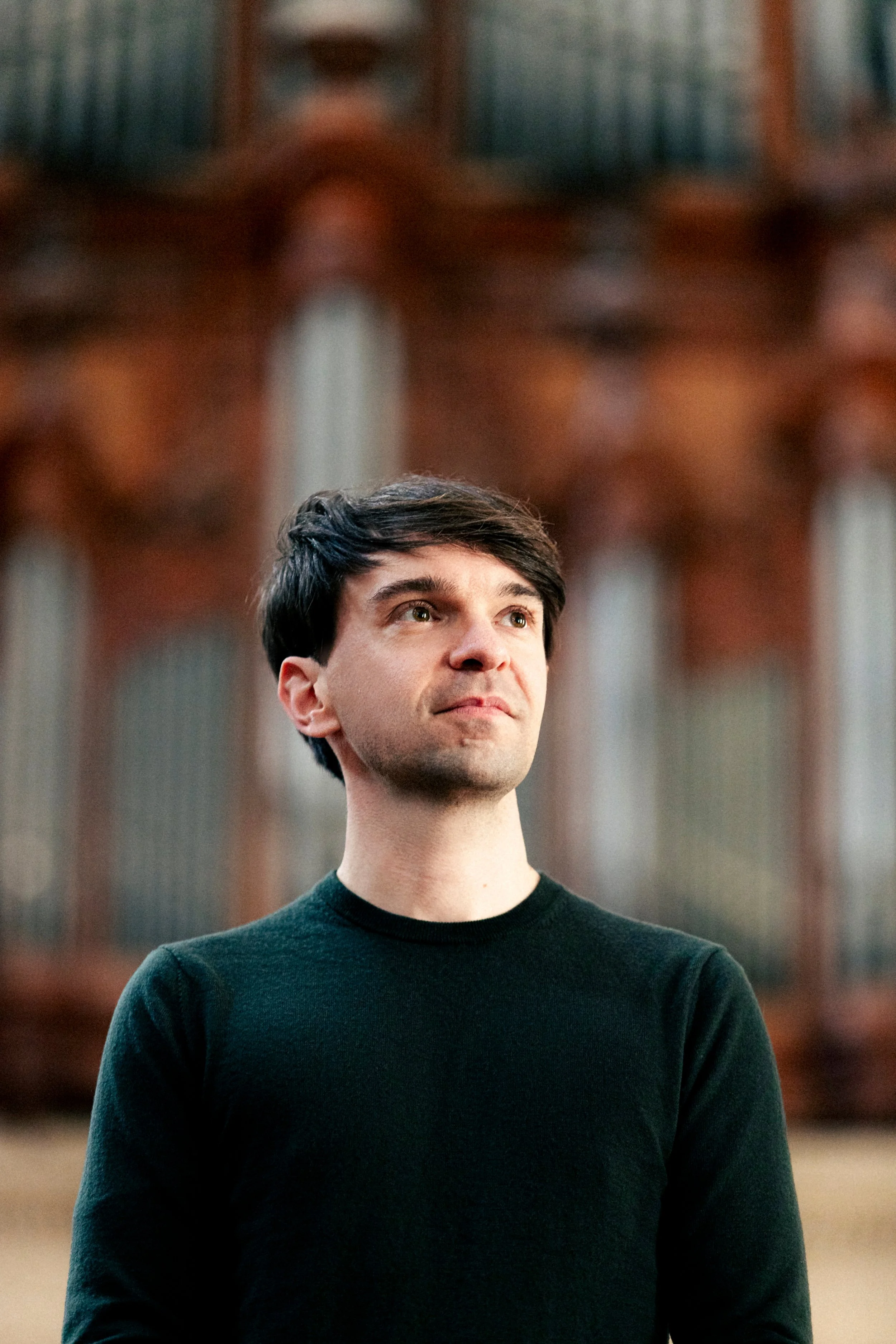 Un jeune homme avec des cheveux bruns, portant un pull noir, se tient dans ce qui semble être une église ou une salle avec une architecture en bois impressionnante en arrière-plan.