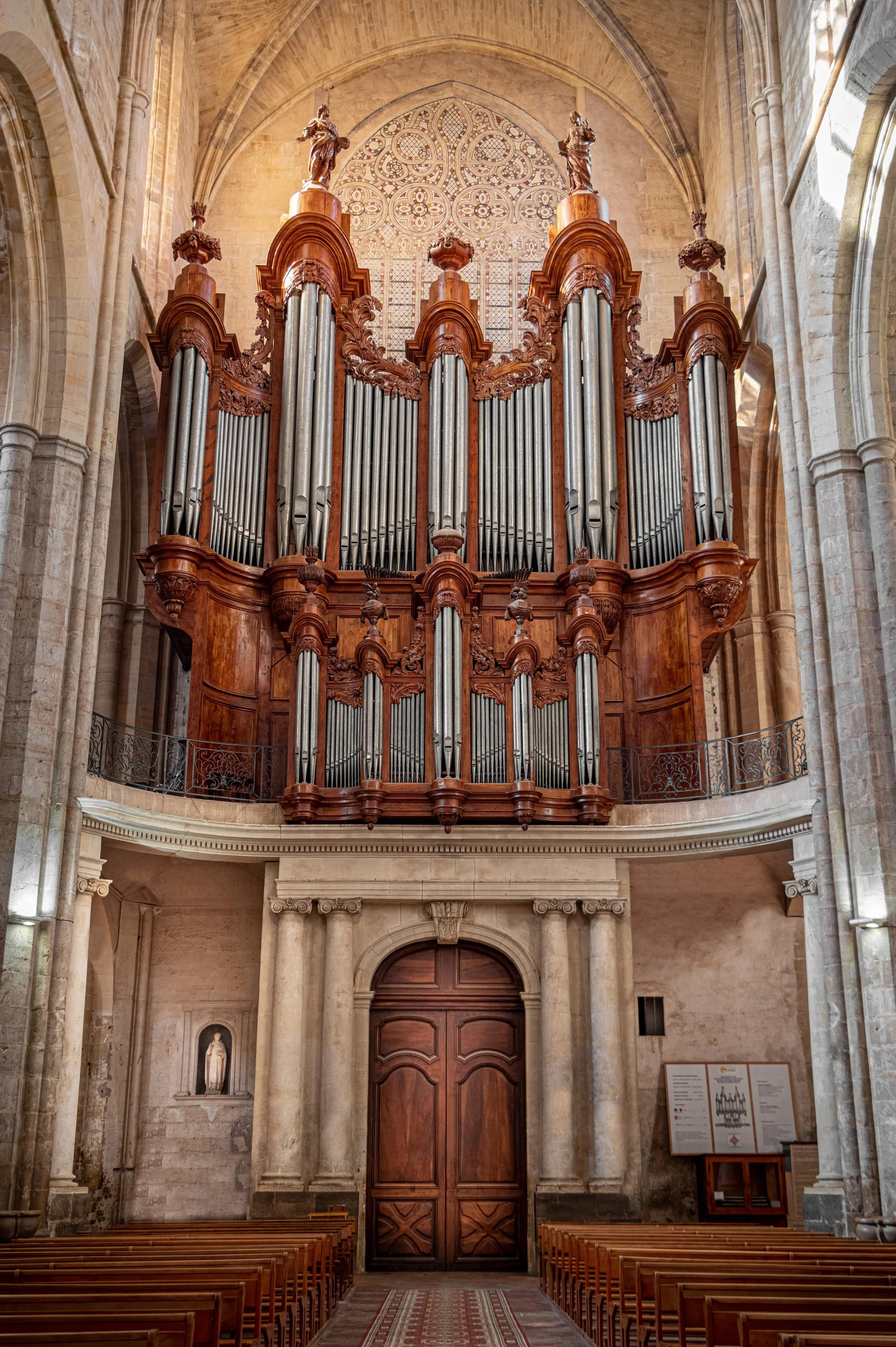 Grand orgue en bois dans une église gothique vue de face, avec des bancs en bois devant et une statue dans une niche à gauche.