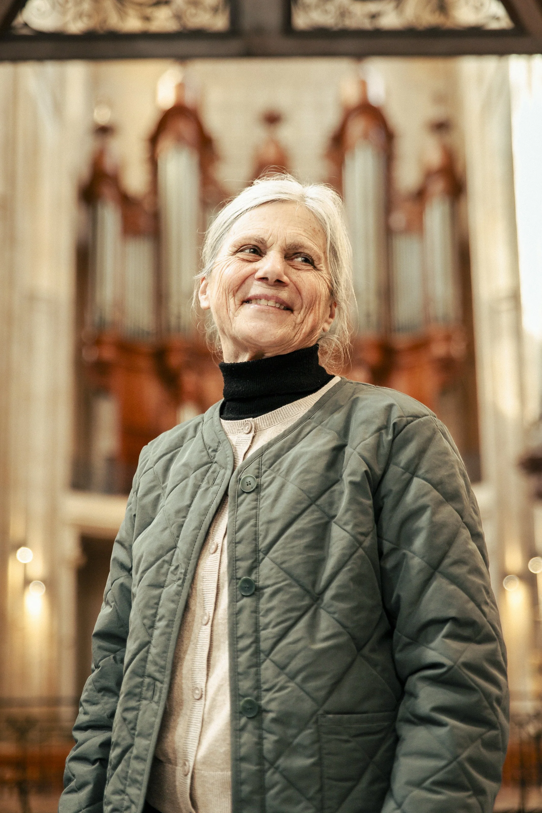 Une femme âgée souriante dans une église avec un orgue en arrière-plan.