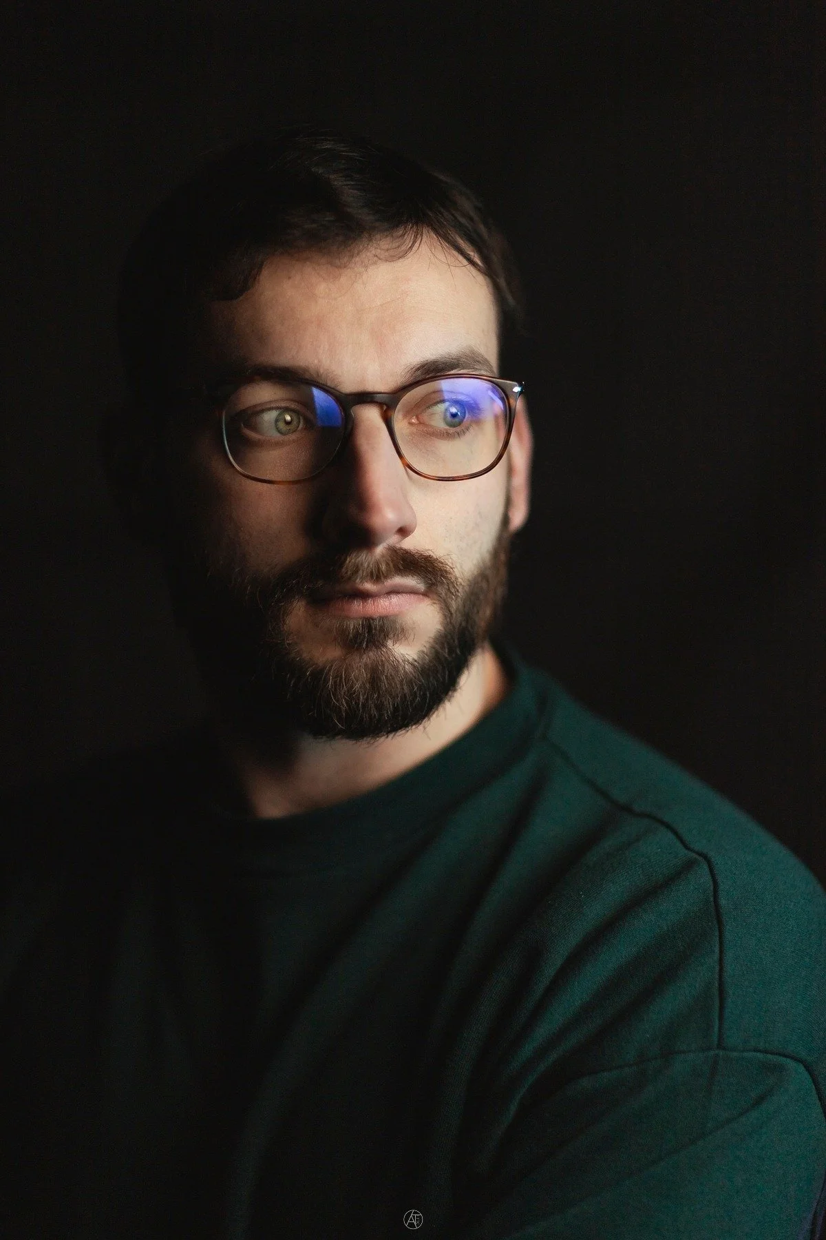 Portrait d'un homme avec des lunettes, une barbe et moustache, portant un t-shirt vert foncé, regardant vers la droite, fond noir.
