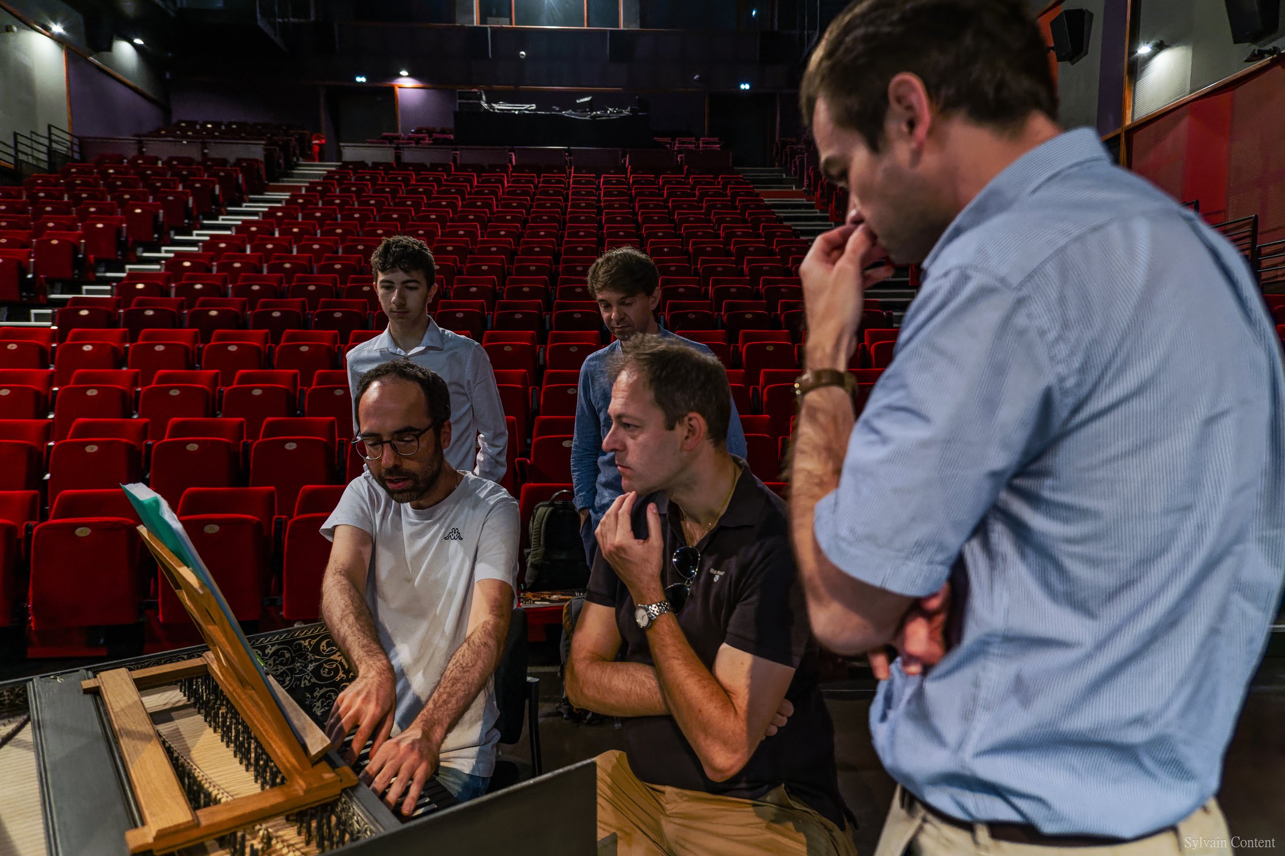 Groupe de cinq personnes dans une salle de théâtre ou de concert avec des sièges rouges, en train de discuter autour d'un piano, portant des vêtements décontractés.