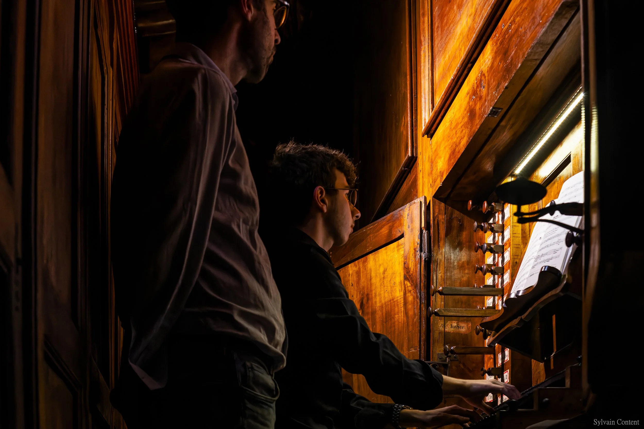 Deux hommes jouent de l'orgue dans une cabine en bois, l'un d'eux est assis et joue du clavier, l'autre se tient derrière lui en observant.