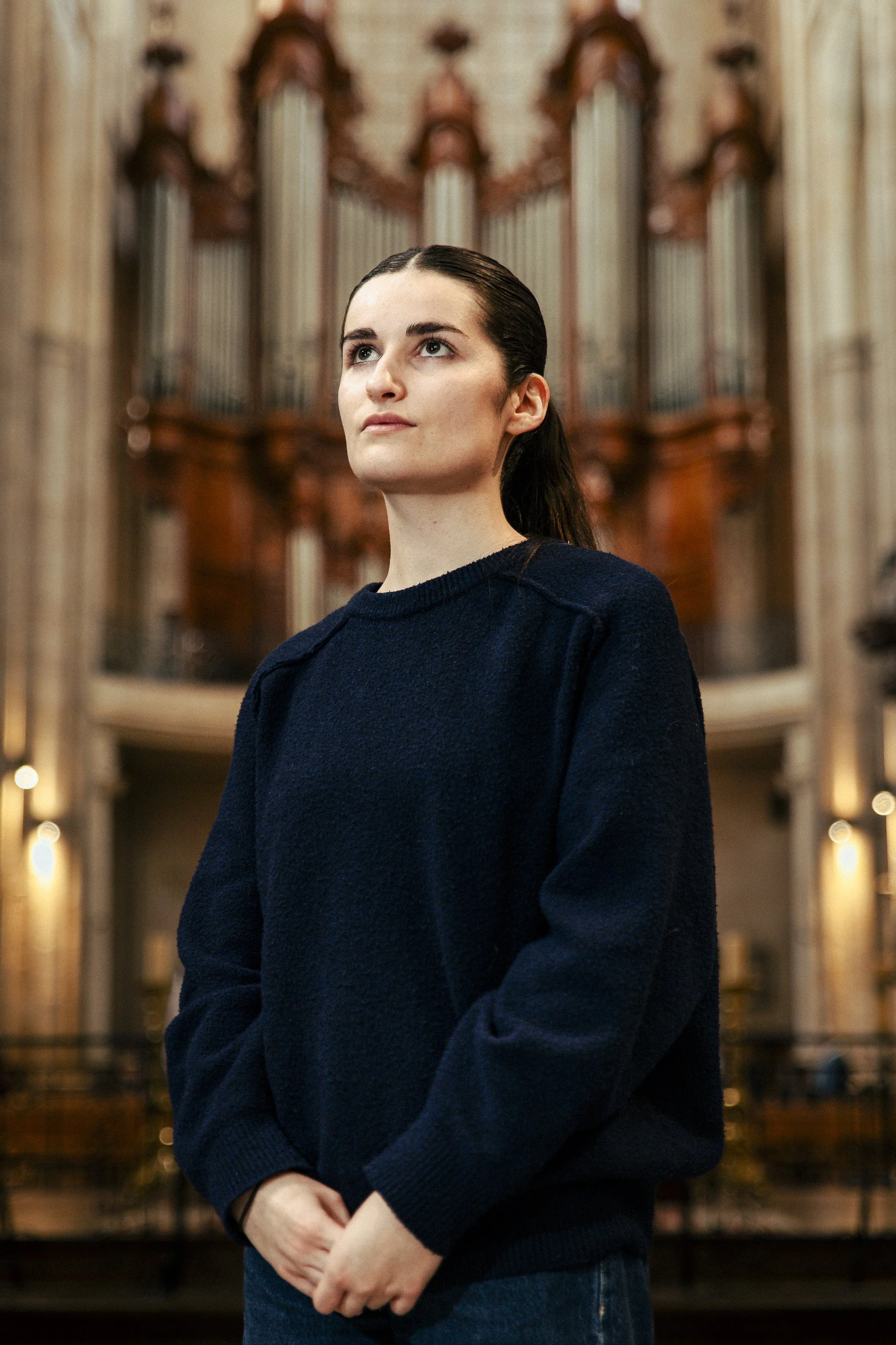 Jeune femme avec une peau claire et cheveux bruns, portant un pull bleu foncé, se tenant dans un lieu avec un orgue en arrière-plan, probablement une église ou une salle de concert.
