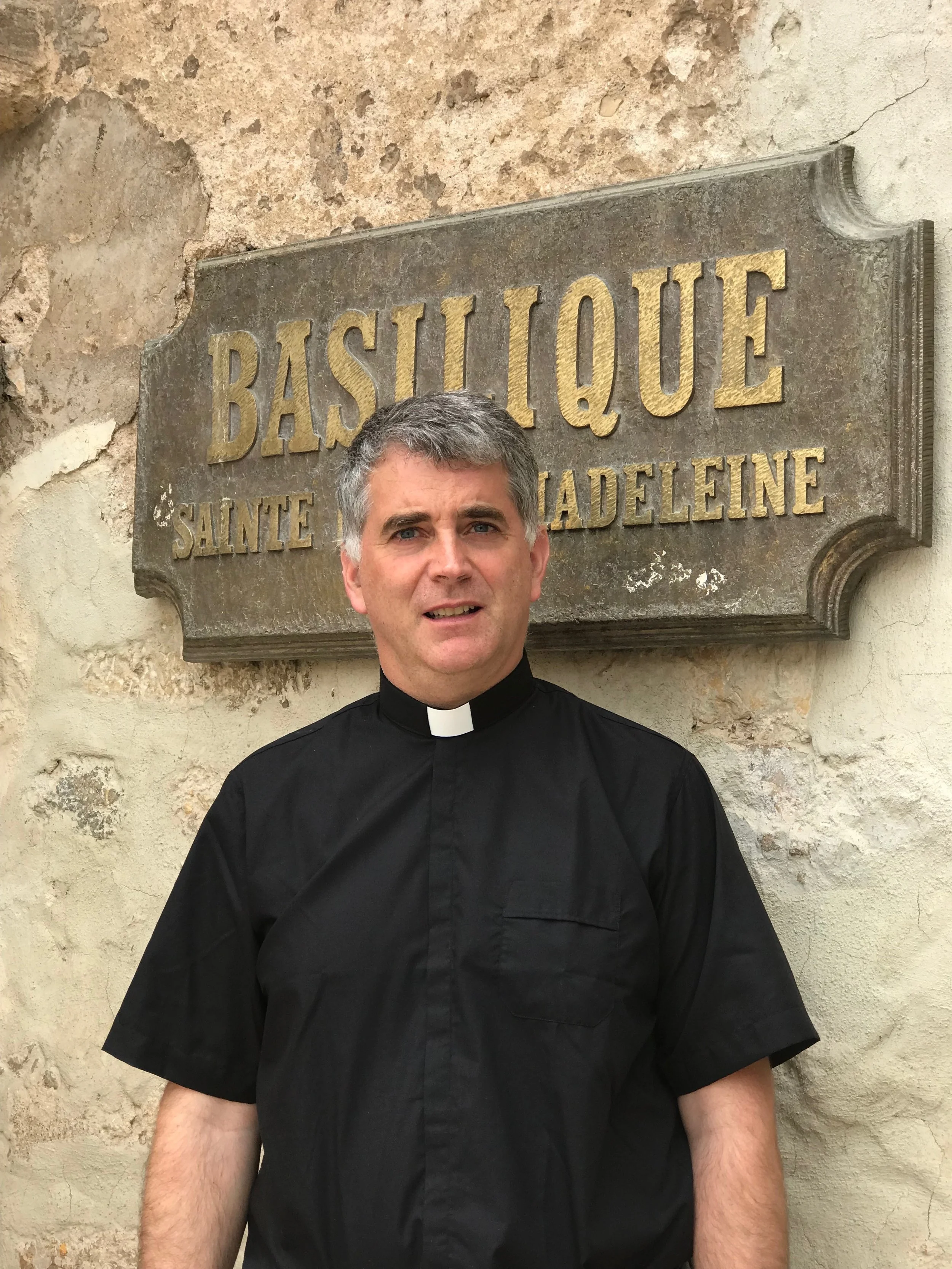 Un prêtre en uniforme noir avec col blanc, se tenant devant une vieille enseigne en pierre portant le mot « BAS-L IQUE » et d'autres mots effacés, devant un mur en pierre.