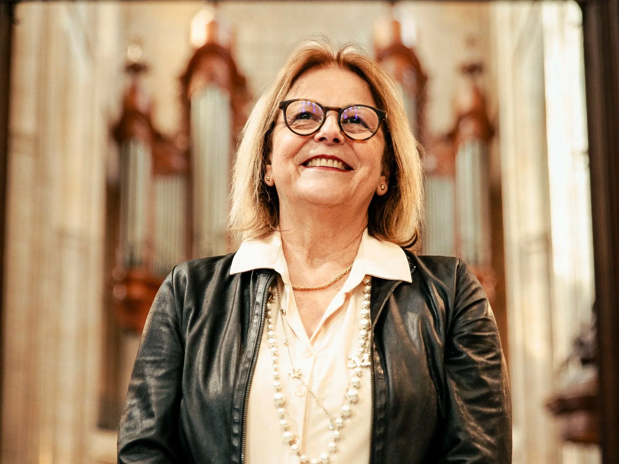 Une femme souriante portant des lunettes, une chemise blanche, une veste en cuir noir, avec des colliers, dans un lieu with des orgues en arrière-plan.