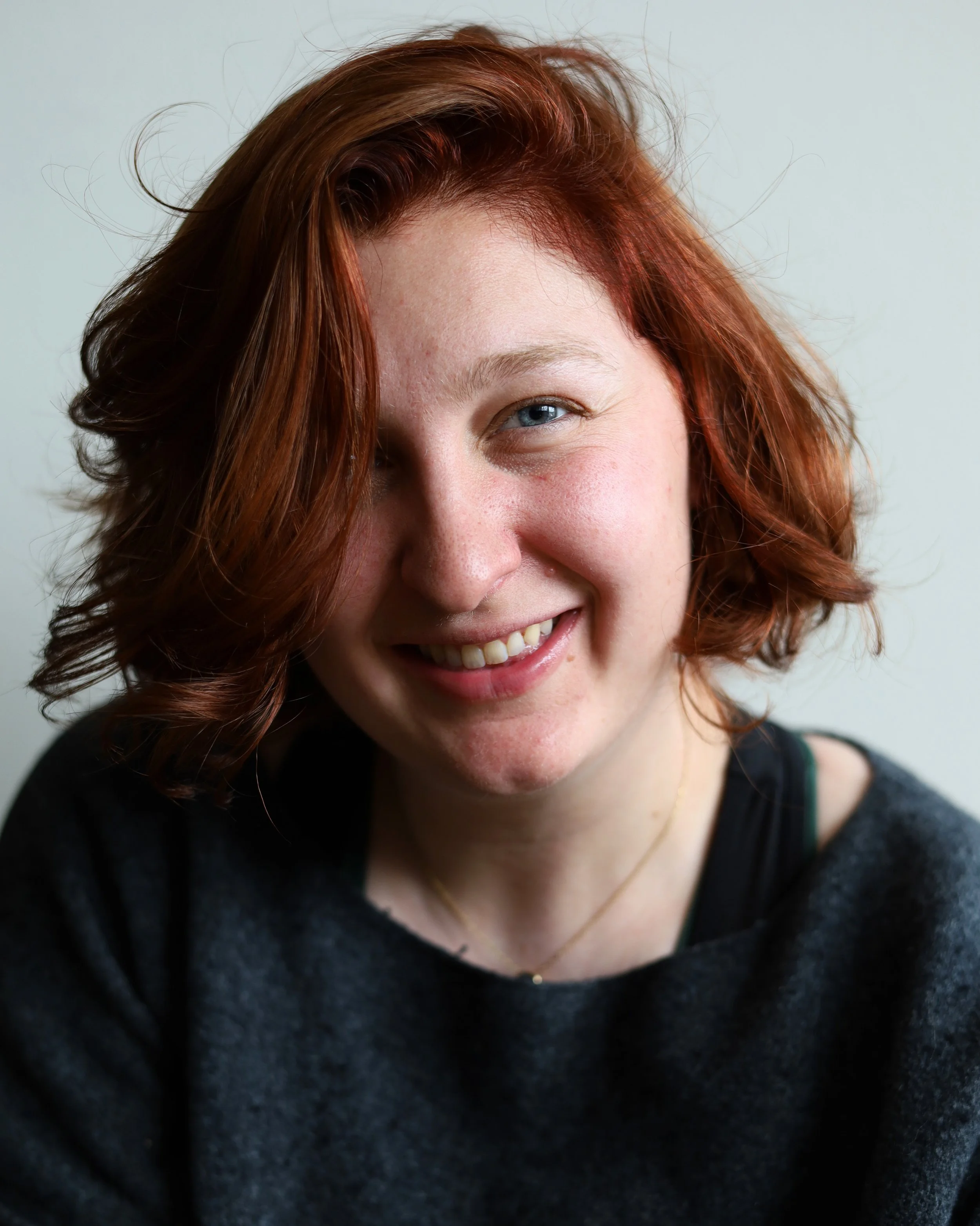 Close-up portrait of a young woman with wavy red hair, blue eyes, and a warm smile, wearing a dark sweater and a necklace, against a plain light background.
