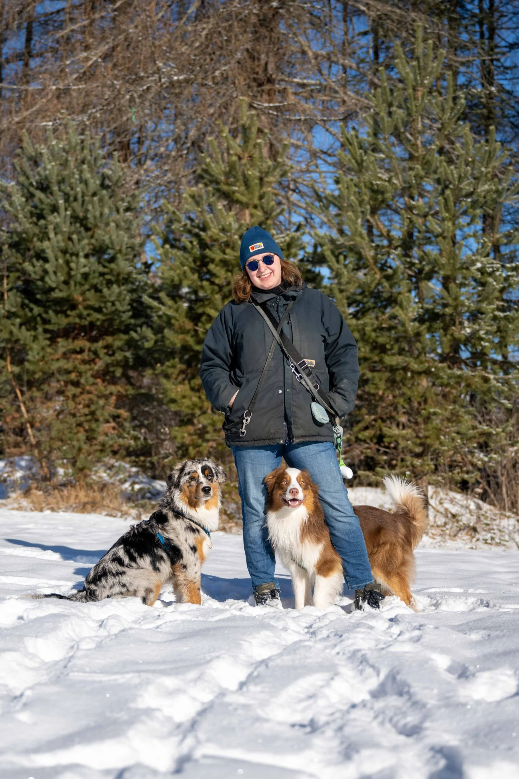 Woman in winter clothing standing on snow with two dogs and a background of pine trees.