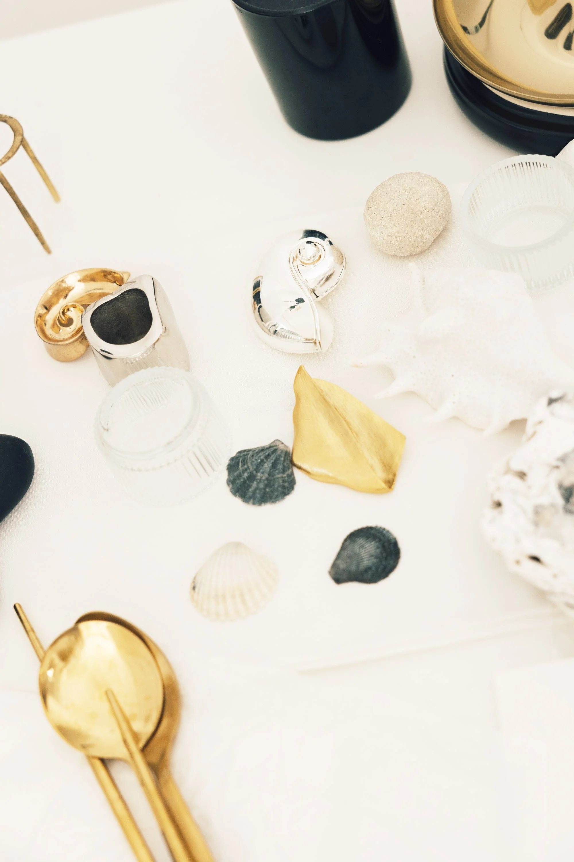 Decorative items on a white surface, including seashells, a gold spoon, a black container, a striped container, and small metallic objects.