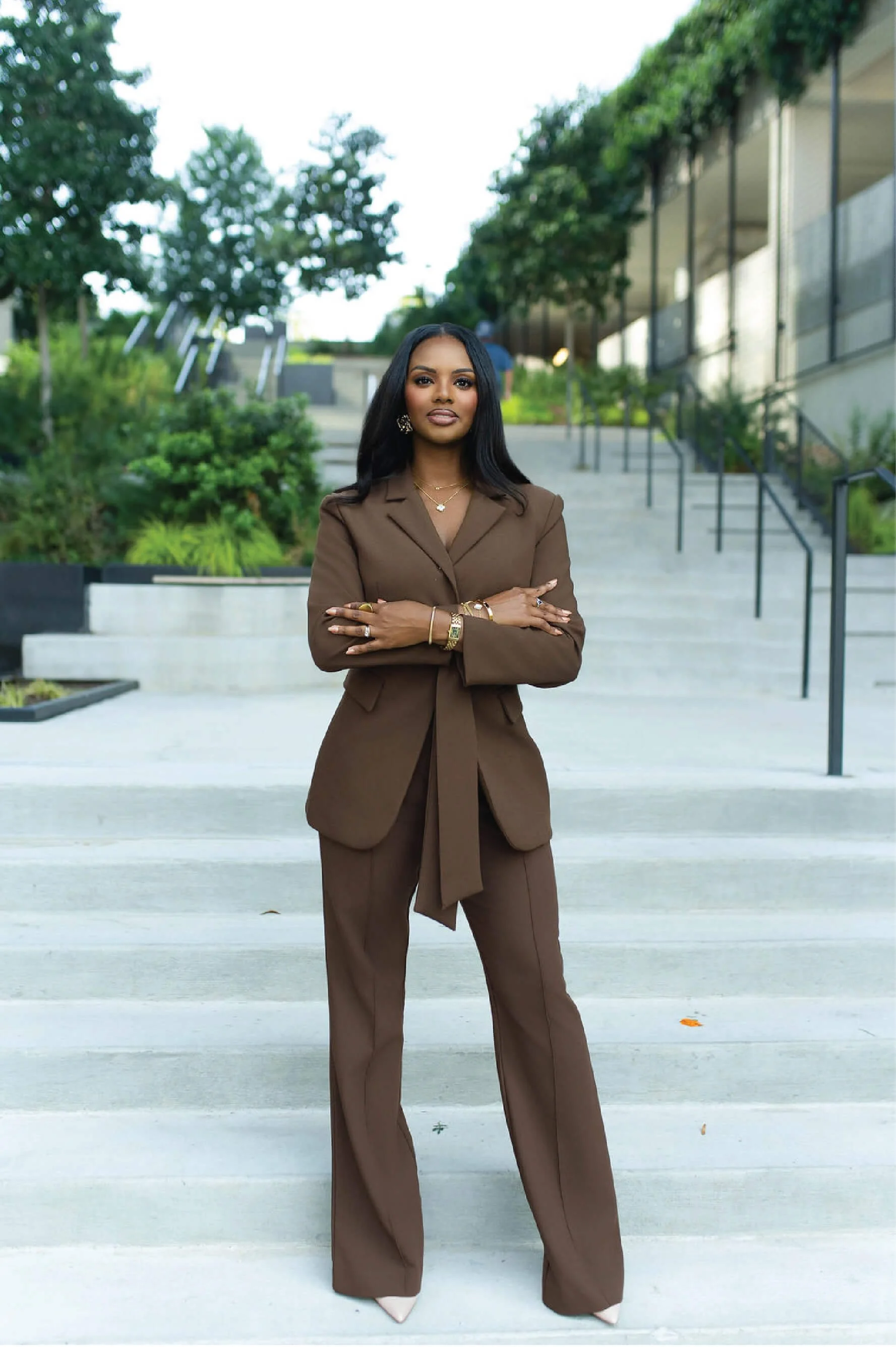A woman in a brown suit standing on outdoor concrete steps with greenery and modern building in the background.