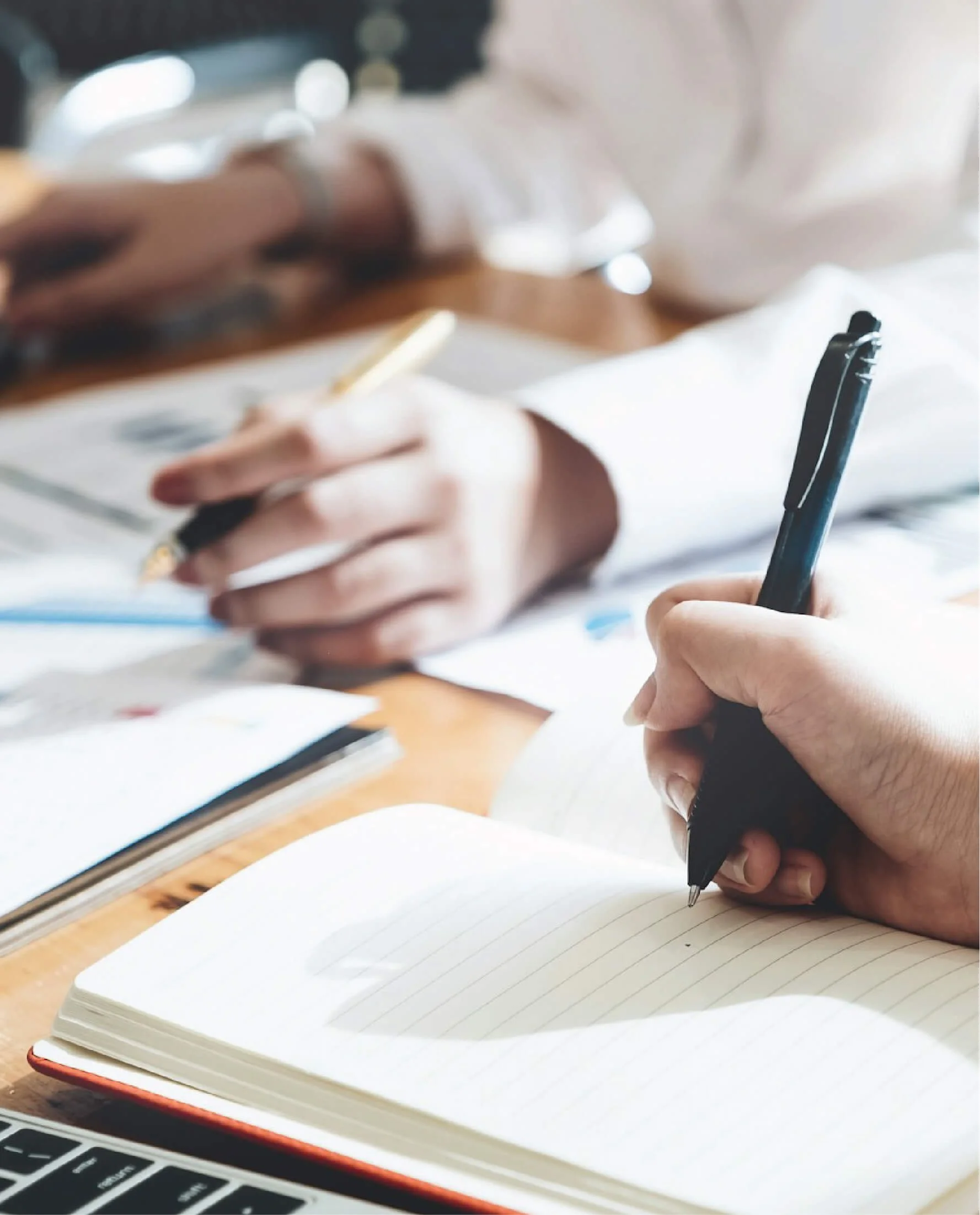 People taking notes with pens on notebooks at a wooden table.