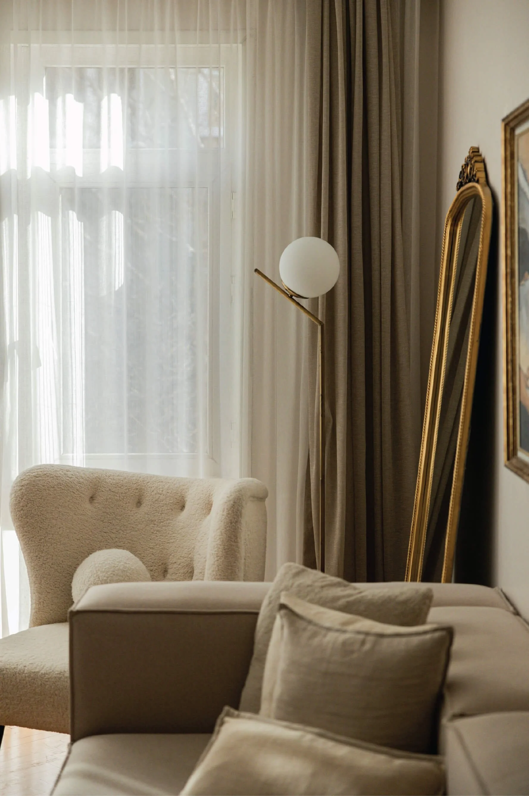 A cozy living room with beige armchairs, a screen door, and gold-framed mirrors, illuminated by natural sunlight.