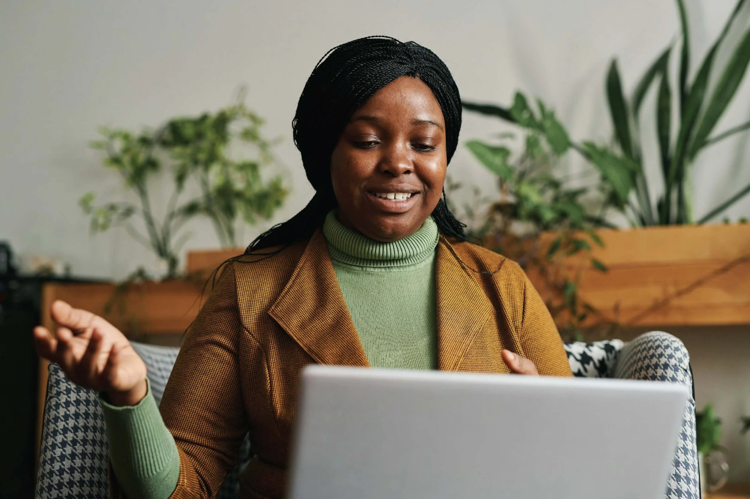 A woman with braids, wearing a green turtleneck and brown blazer, sitting at a table in front of a laptop, smiling and gesturing with her hand.