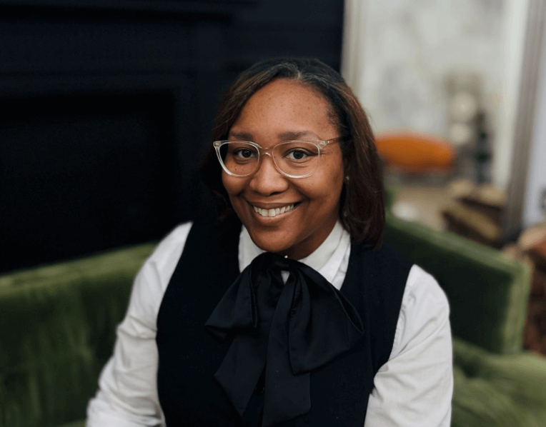 A woman smiling, wearing glasses, a white shirt, and a black vest with a bow, sitting on a green sofa.