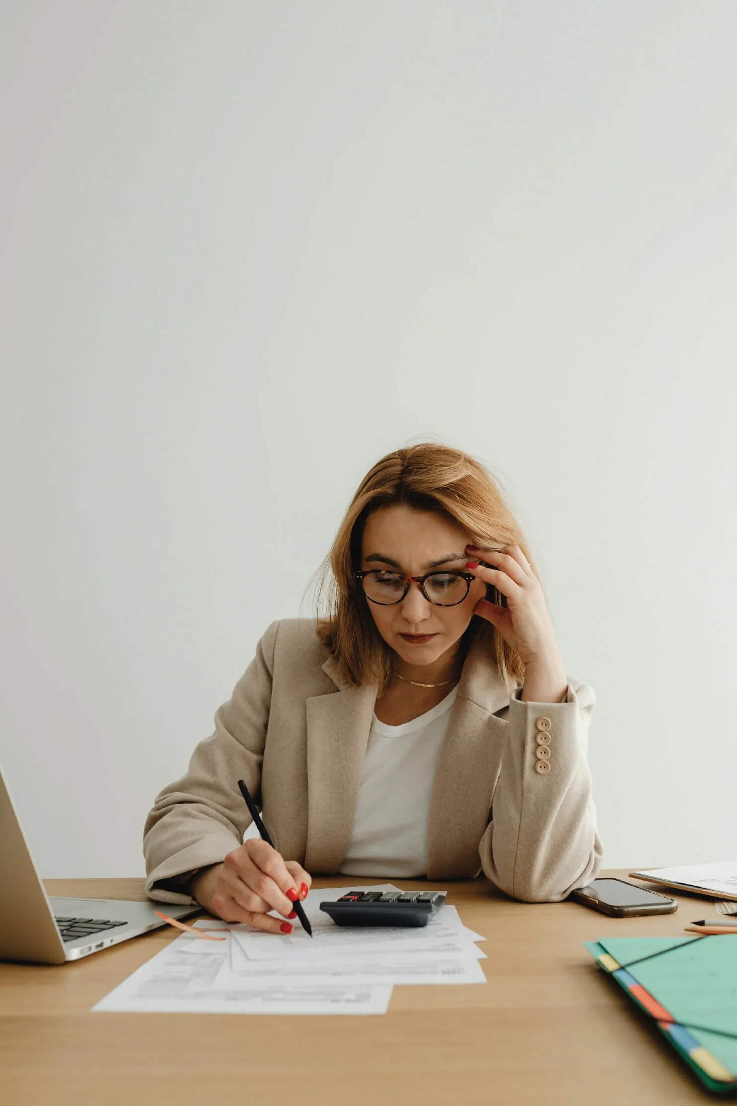 A woman with red hair and glasses working at a wooden desk, reviewing documents with a calculator, laptop, and folders.