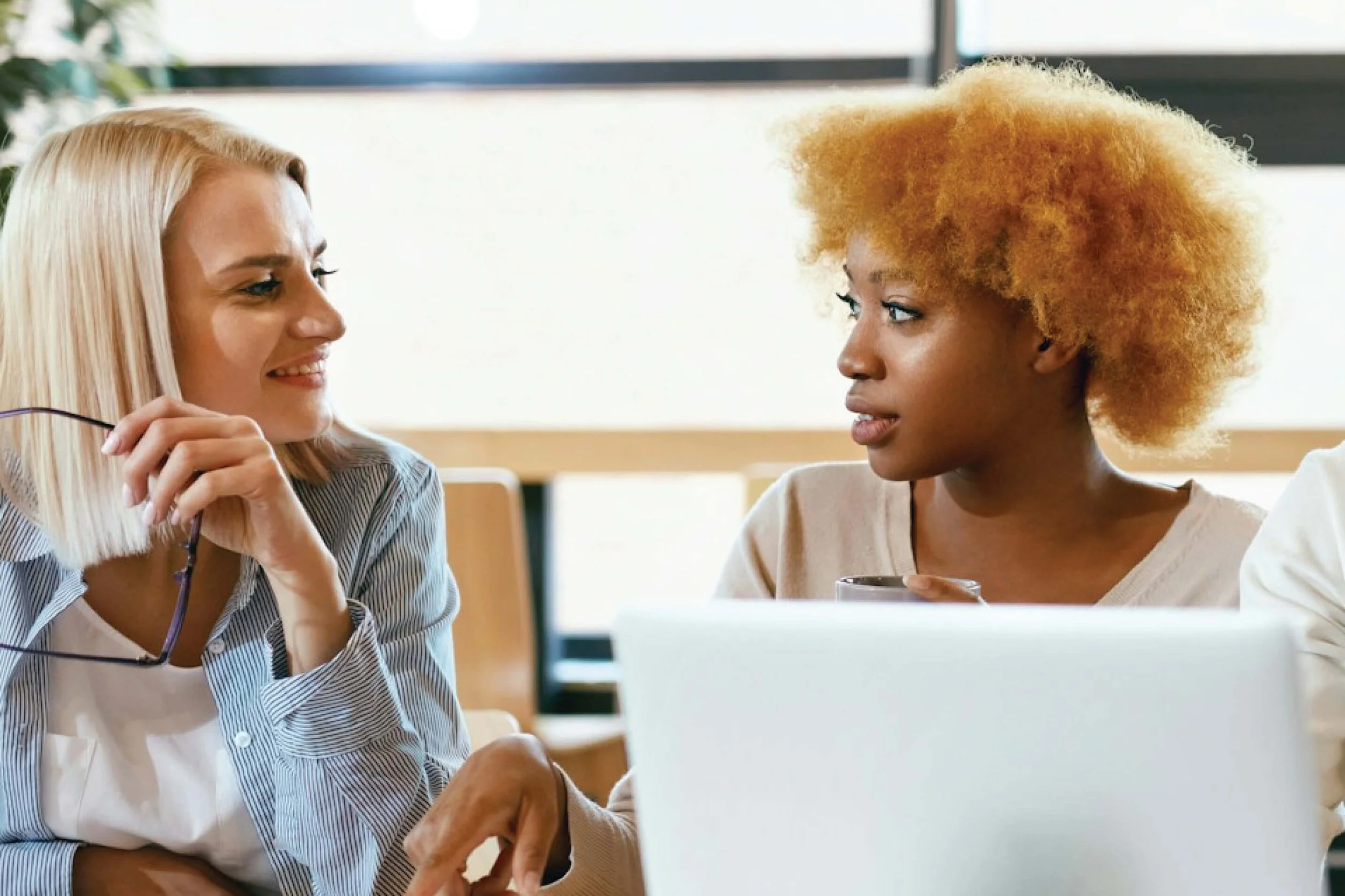 Two women engaged in conversation, one holding eyeglasses and the other with a laptop in front of her, in a well-lit room.