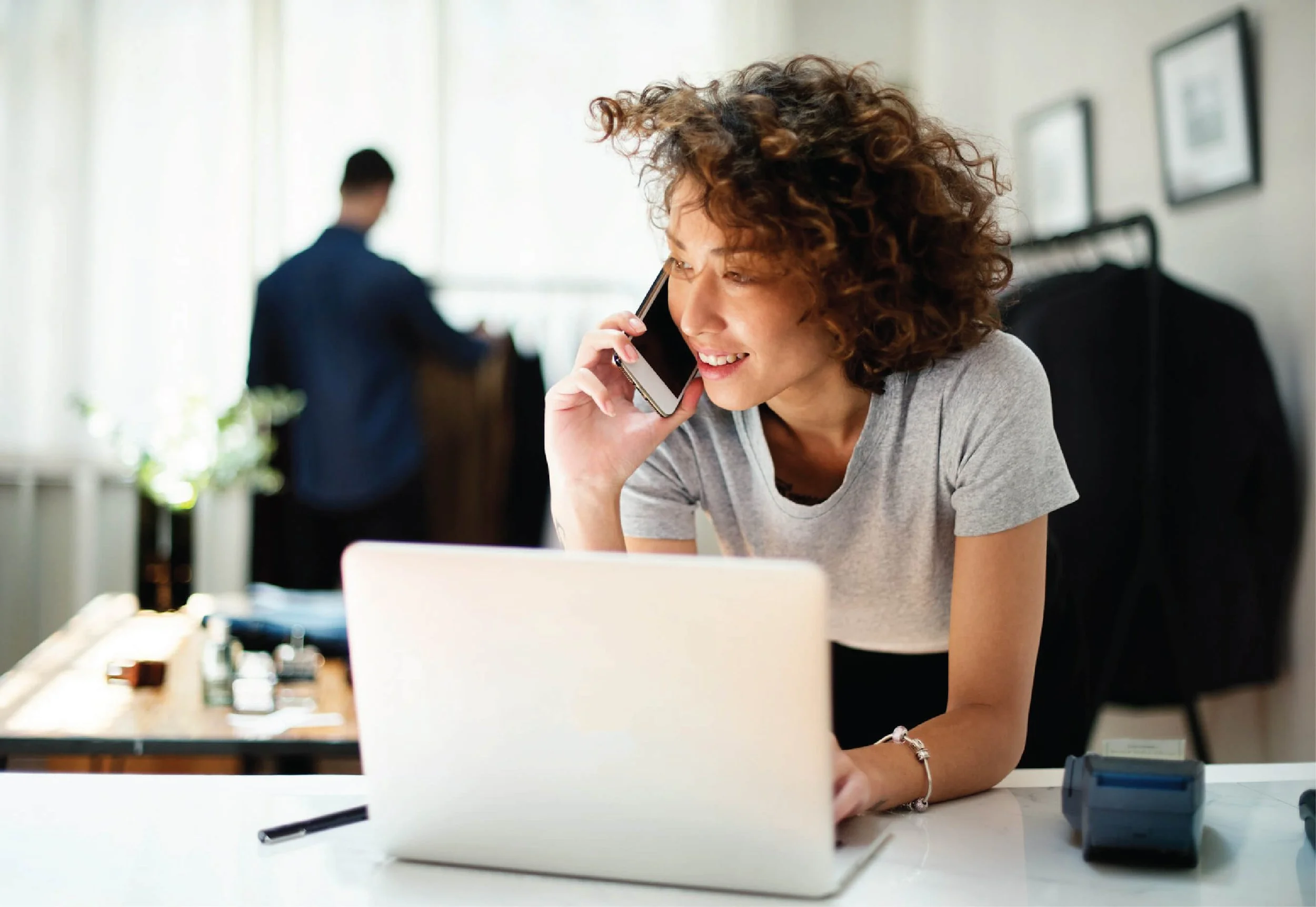 A woman with curly hair talking on her cellphone while working on her laptop at a desk in an office.