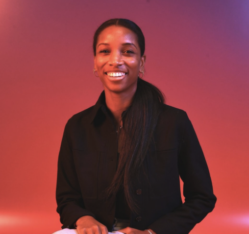 A woman with long dark hair, wearing a black shirt, smiling in front of a pink and purple gradient background.
