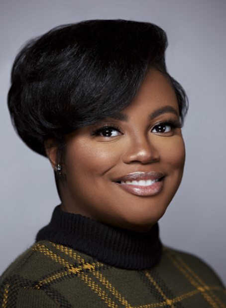 A professional woman with short black hair, wearing a black turtleneck and a green plaid jacket, smiling at the camera.