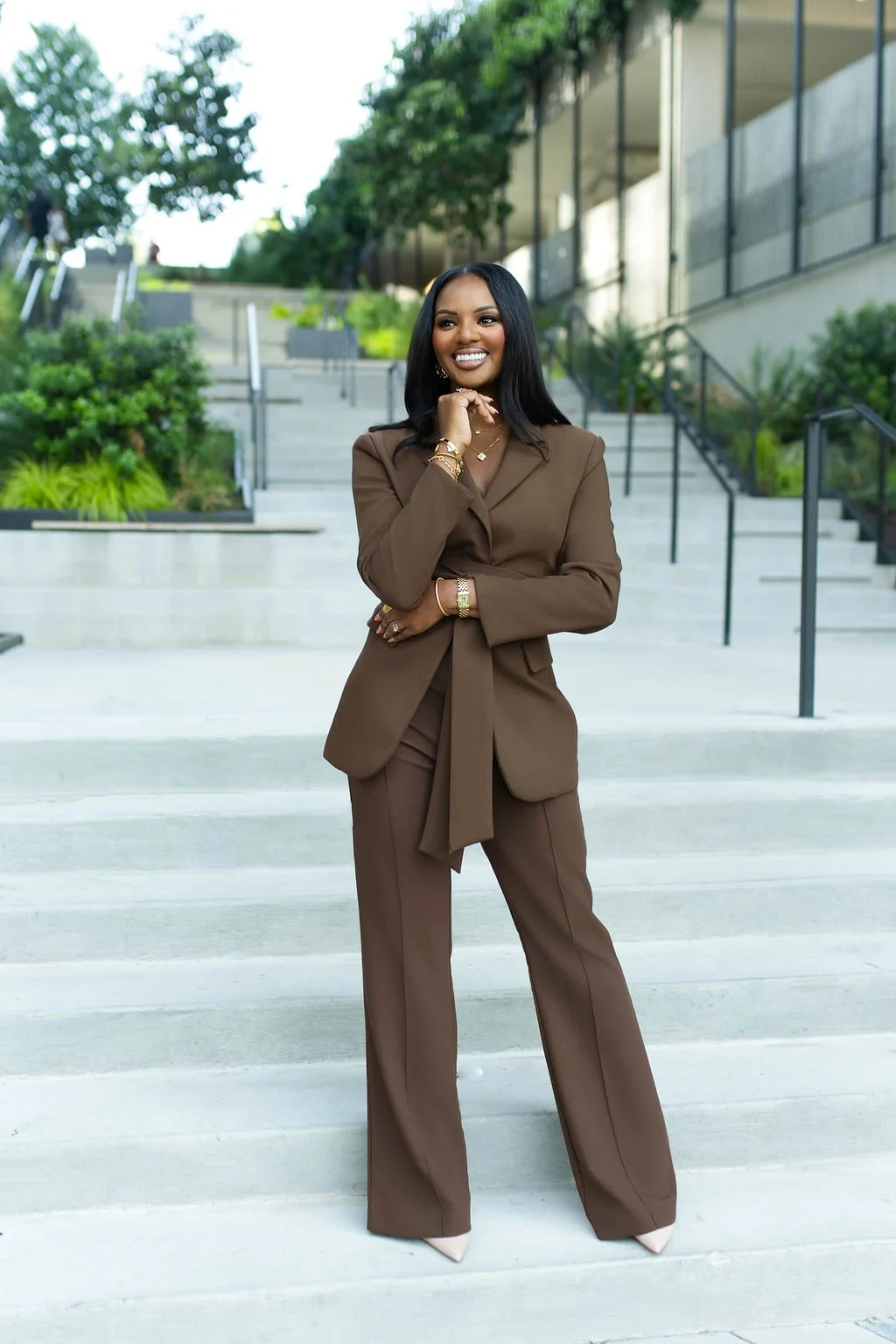 A confident woman in a brown suit standing on outdoor stairs with greenery and modern building in the background, smiling and posing with her hand near her chin.