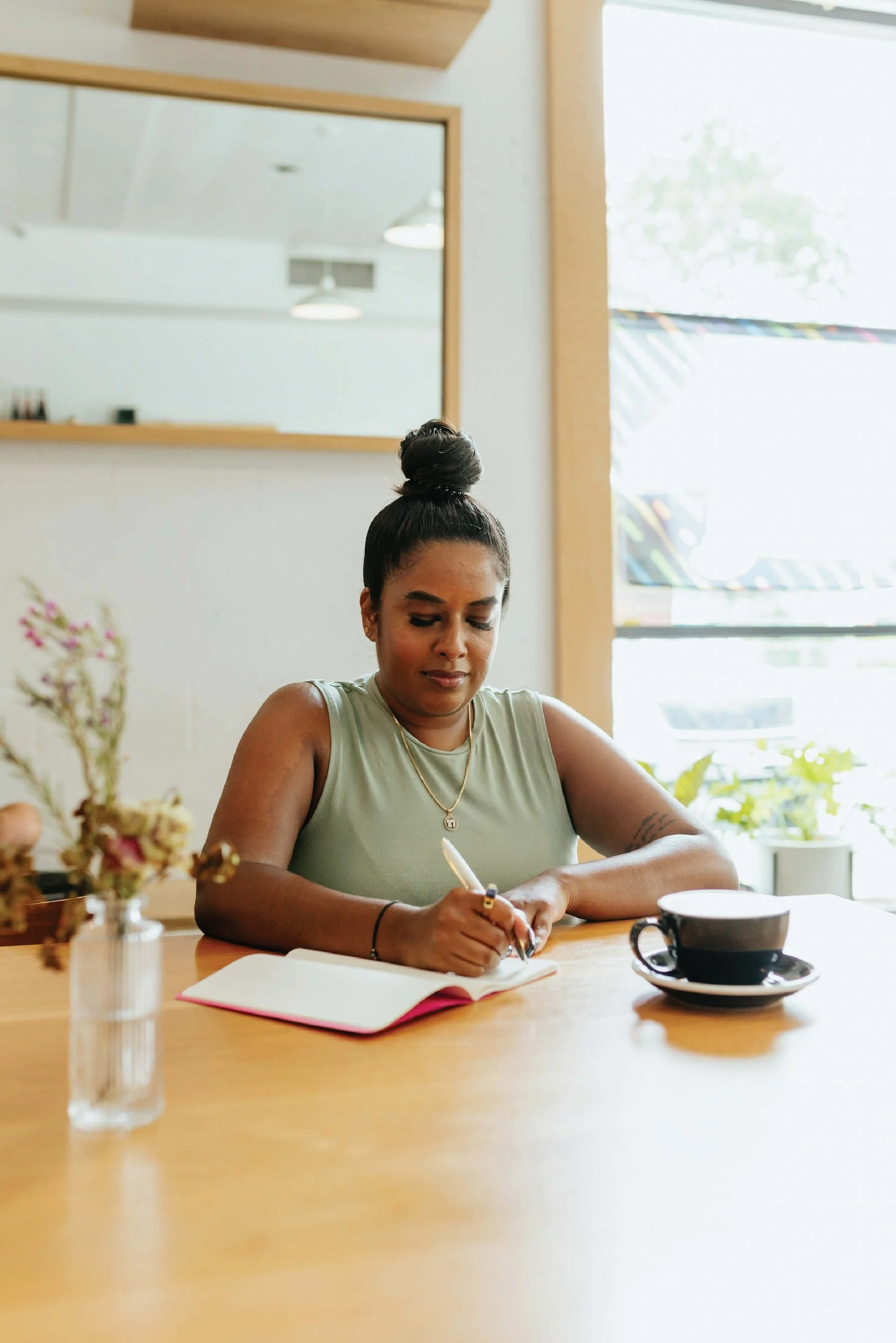 A woman with dark hair styled in a bun, wearing a green sleeveless top and jewelry, sitting at a wooden table writing in a notebook with a pen, a black coffee cup and saucer, and a vase of flowers in a bright cafe with large windows.
