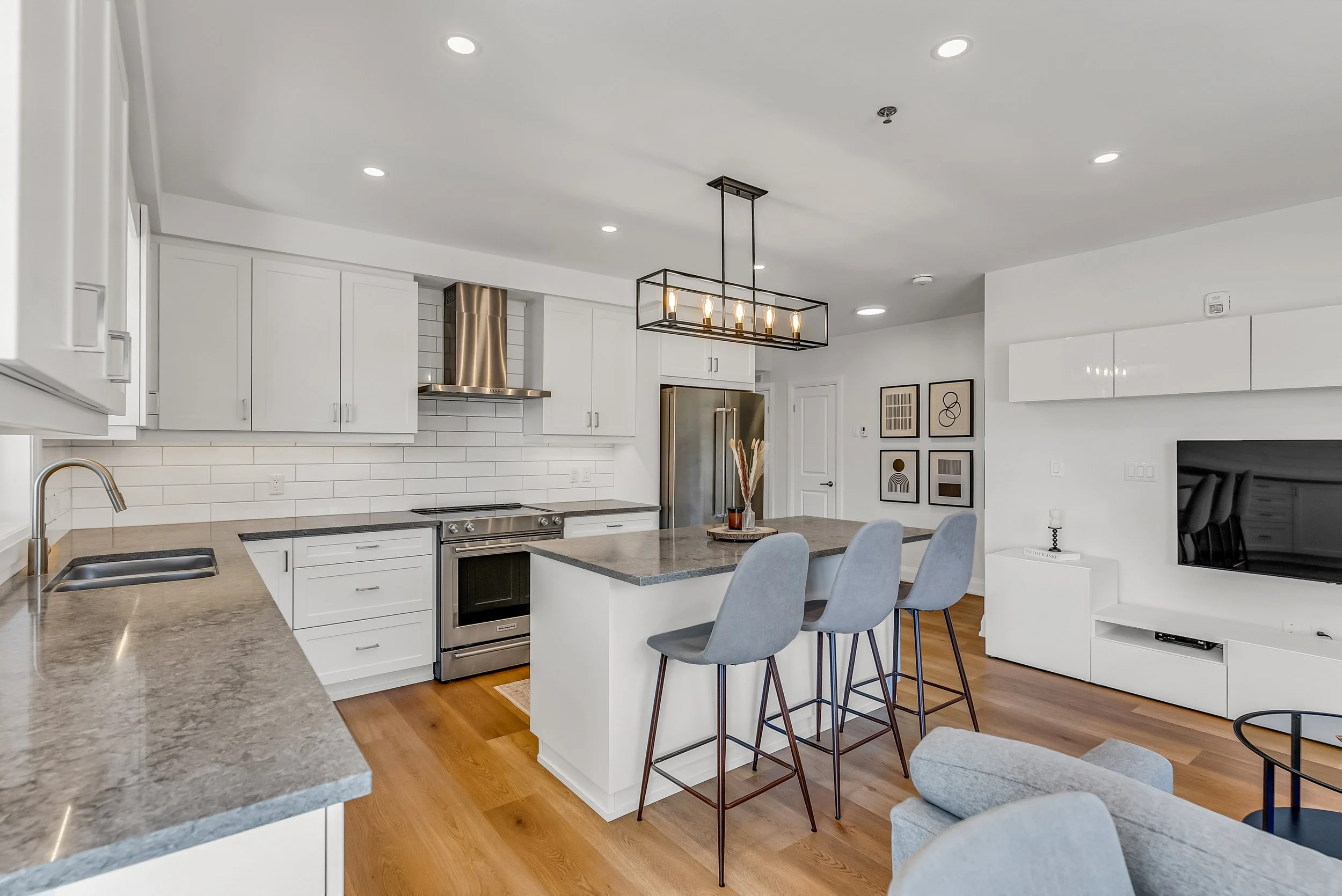 Modern kitchen with white cabinets, gray countertops, stainless steel appliances, and a kitchen island with three gray bar stools. There is a black rectangular chandelier above the island, white tiled backsplash, and wood flooring. In the background, there are framed abstract art pieces and a built-in white entertainment unit with a television.