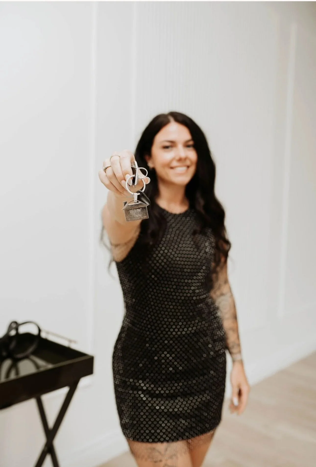 A woman in a black, sequin dress standing in a room with white walls, smiling and holding up a set of keys towards the camera.