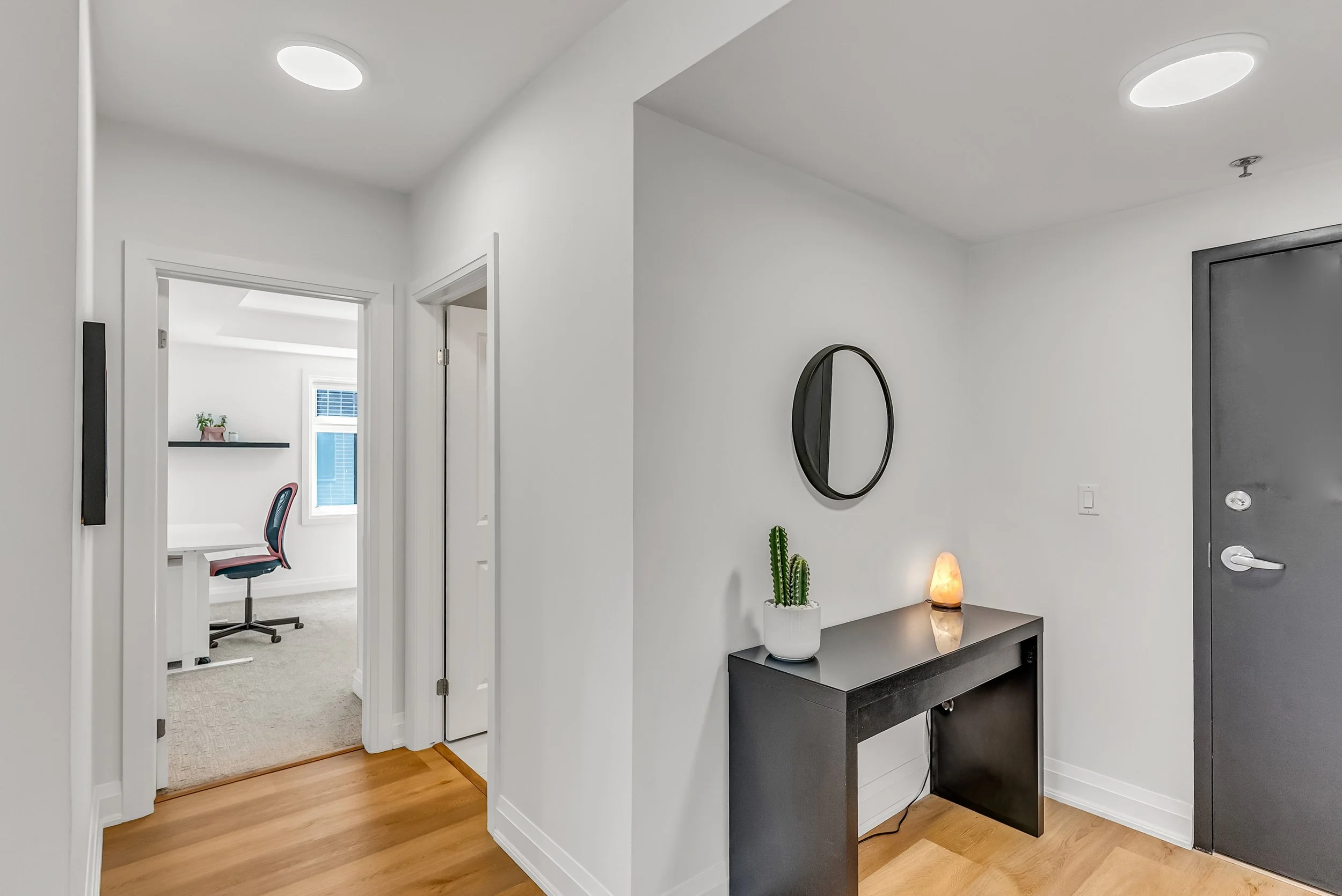 Entryway with a black console table, potted cactus, salt lamp, round mirror, white walls, and a gray door. An open doorway reveals a home office with a desk, pink and black office chair, window, and a floating shelf with a plant.