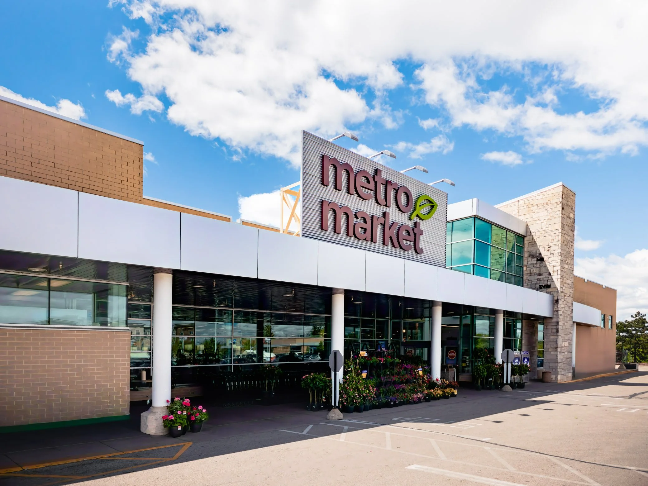 Exterior view of a modern shopping center with a sign that reads 'market' under a blue sky with white clouds. There are tables and chairs outside with potted flowers.