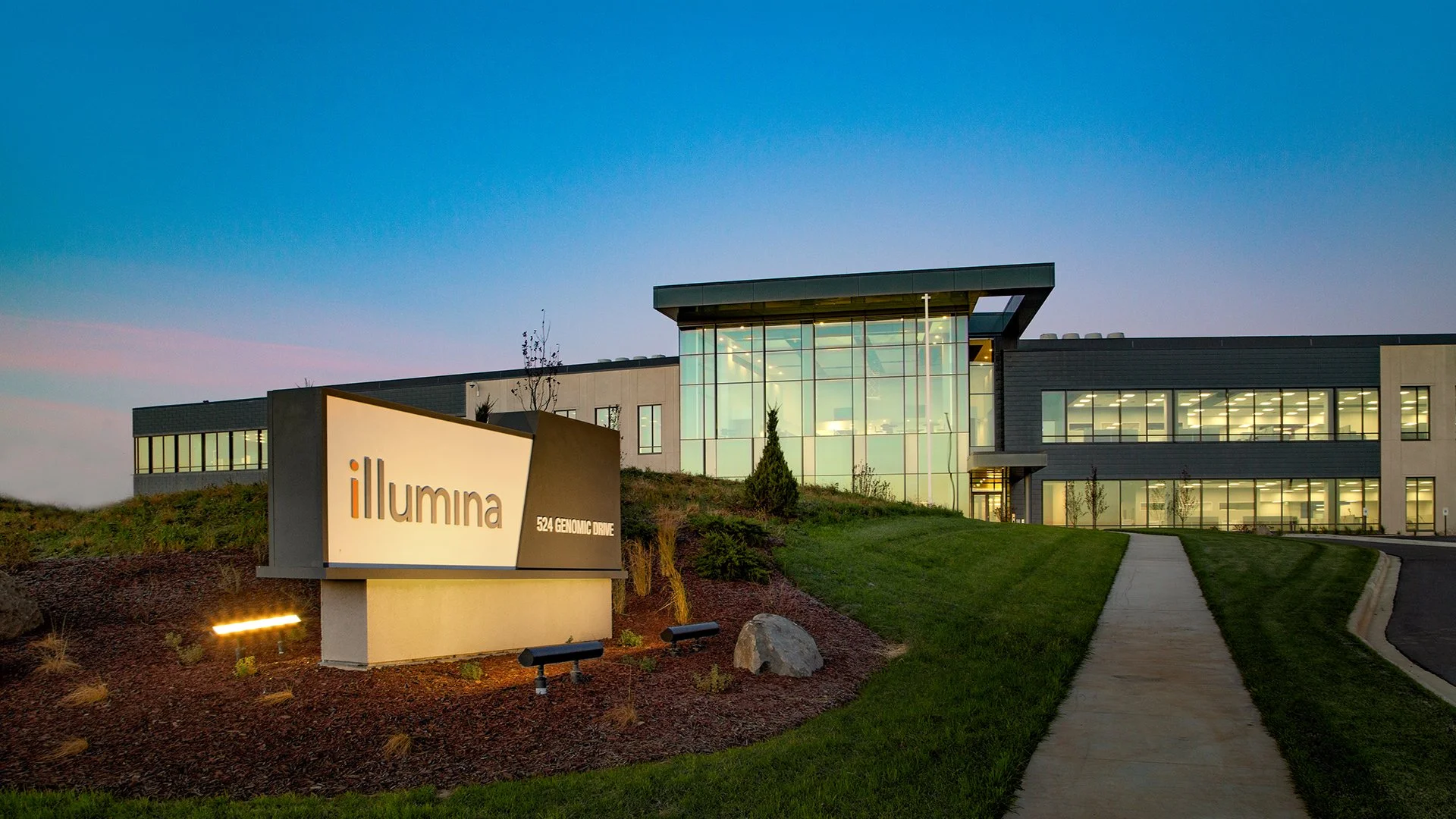 Modern office building with large glass windows, illuminated from inside at dusk. A sign in front reads "Illumina, 524 Genomic Drive." The building has a landscaped area with small trees, shrubs, and a pathway leading to the entrance.