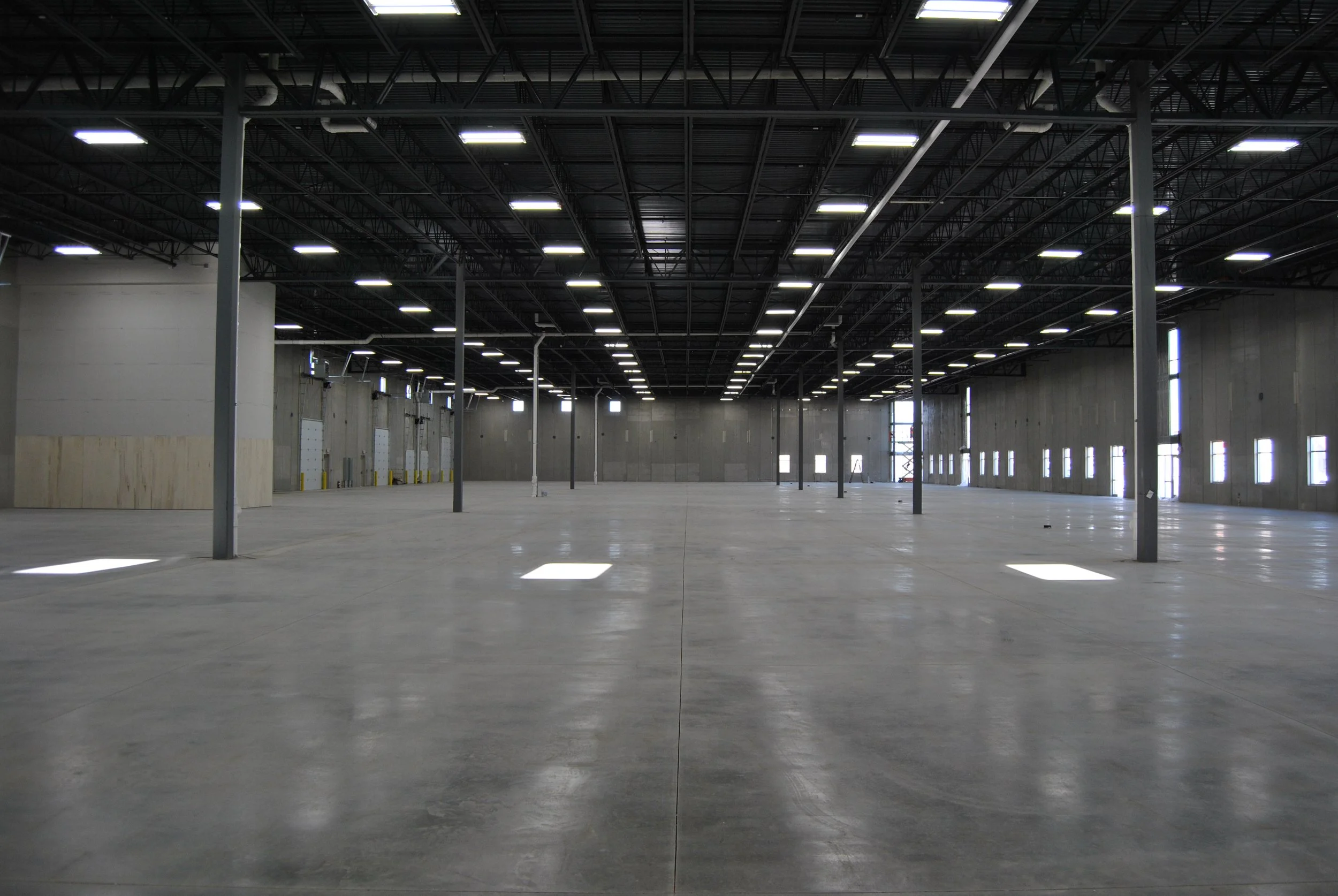 A large, empty warehouse with a high ceiling, exposed metal framework, concrete floor, and evenly spaced overhead lights. The warehouse has unadorned concrete walls and small windows near the ceiling, with no furniture or equipment visible.