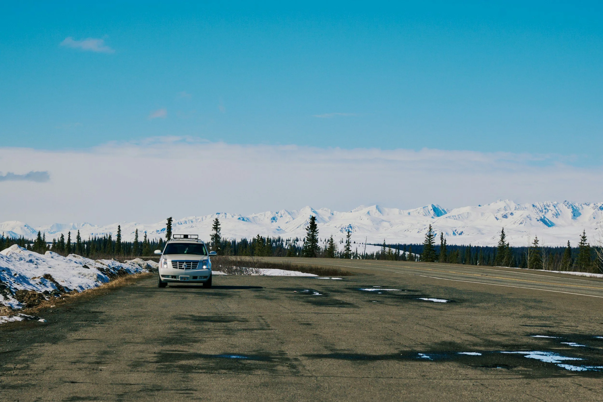 A silver car parked on the side of a mountain road with snow patches, surrounded by pine trees, with snow-covered mountains in the background under a blue sky.