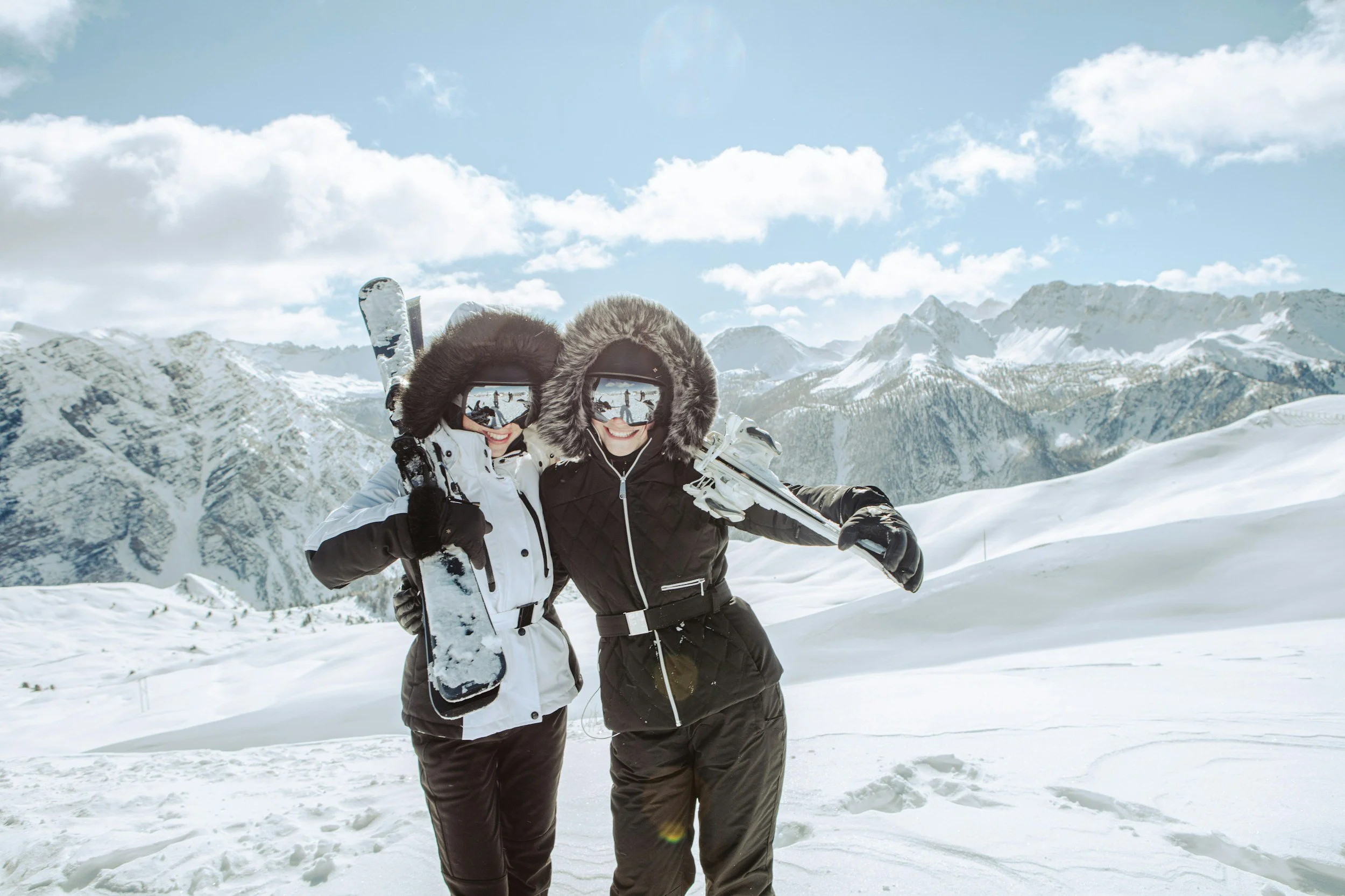 Two women in winter clothing with fur-lined hoods and reflective ski goggles posing with snowboards and skis in a snowy mountain landscape.