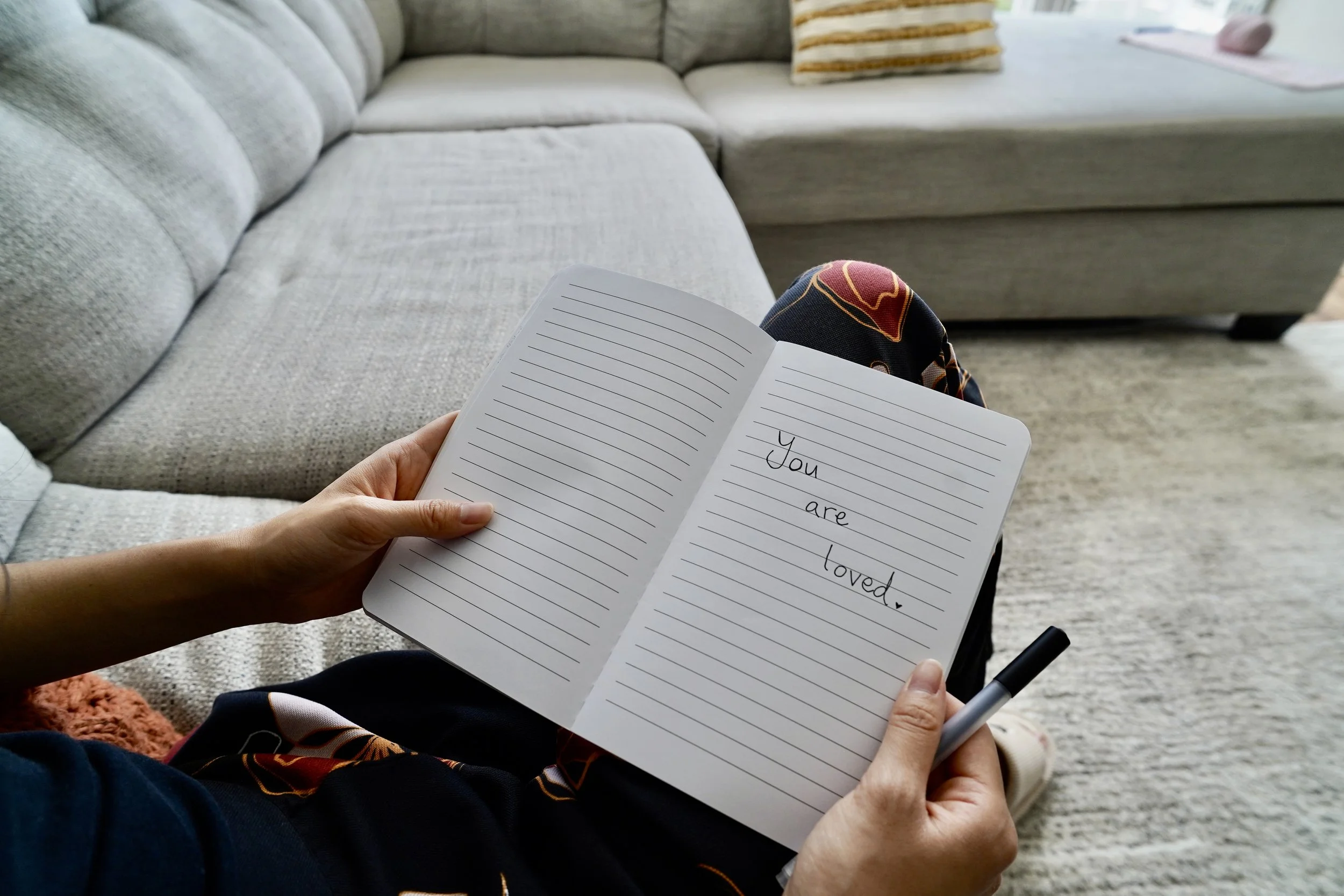 Person holding open notebook with handwritten message, 'You are loved,' in a living room with a gray sofa and patterned pillow.
