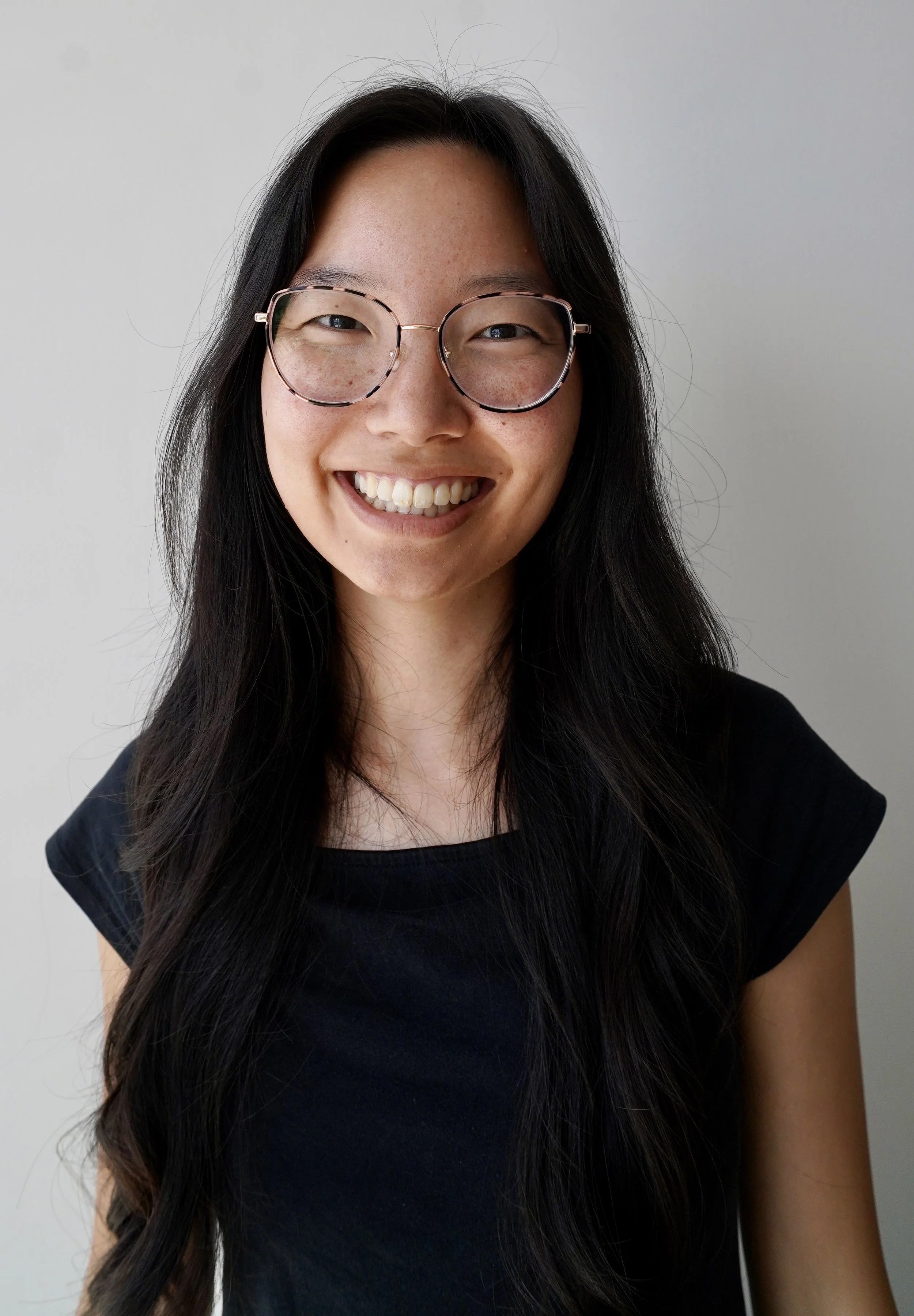 A woman with long black hair, glasses, and a black top smiling in front of a plain light-colored background.