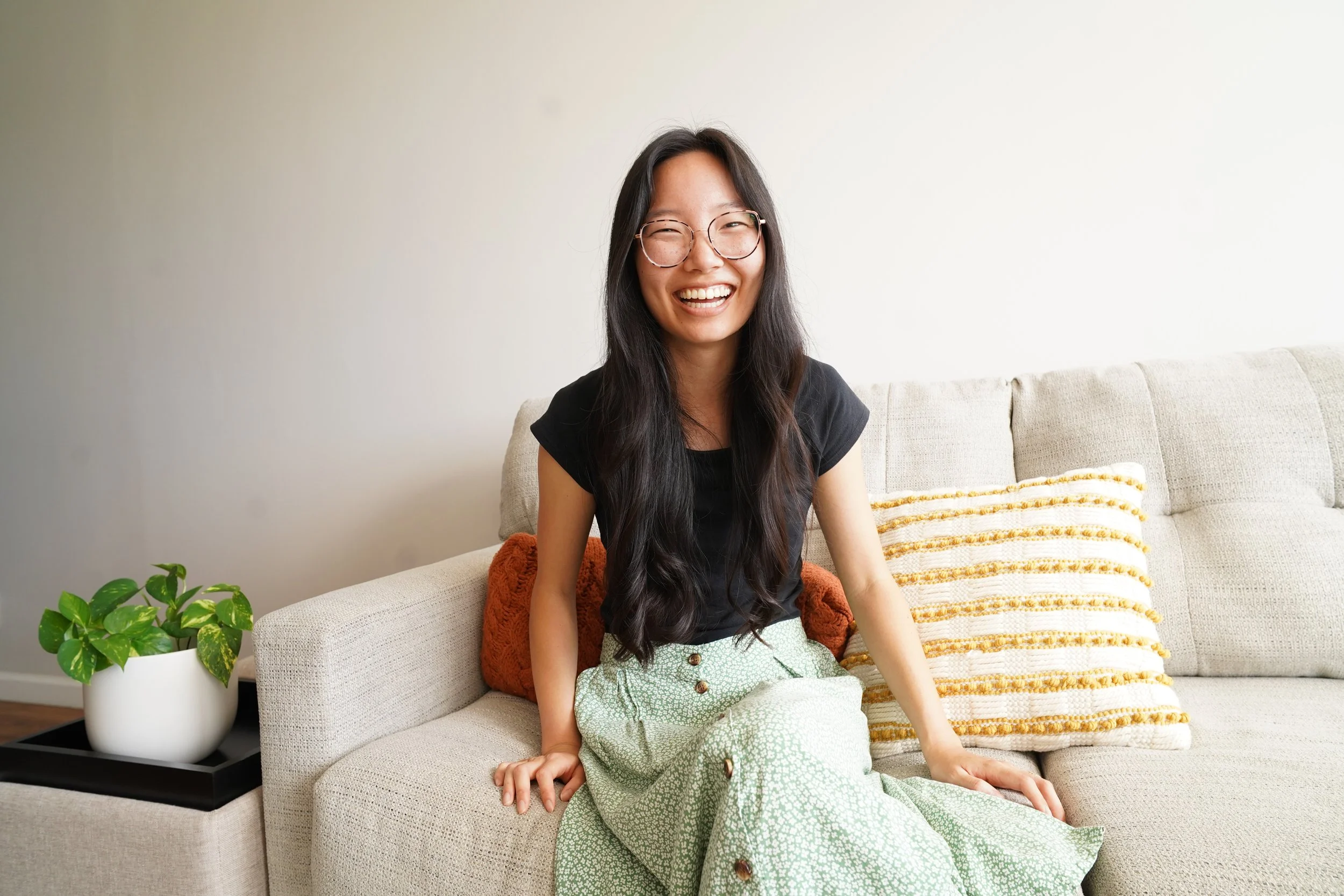 A woman with long dark hair, glasses, and a black top sitting on a beige sofa, smiling. There is a plant on a side table and patterned pillows on the sofa.