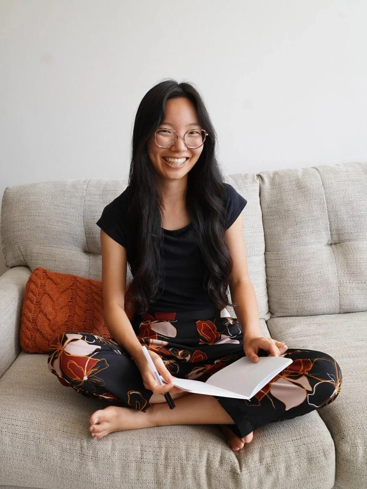 A woman sitting on a beige couch, smiling, wearing glasses, a black t-shirt, and floral patterned pants, holding a notebook and pen, with an orange knit pillow behind her.
