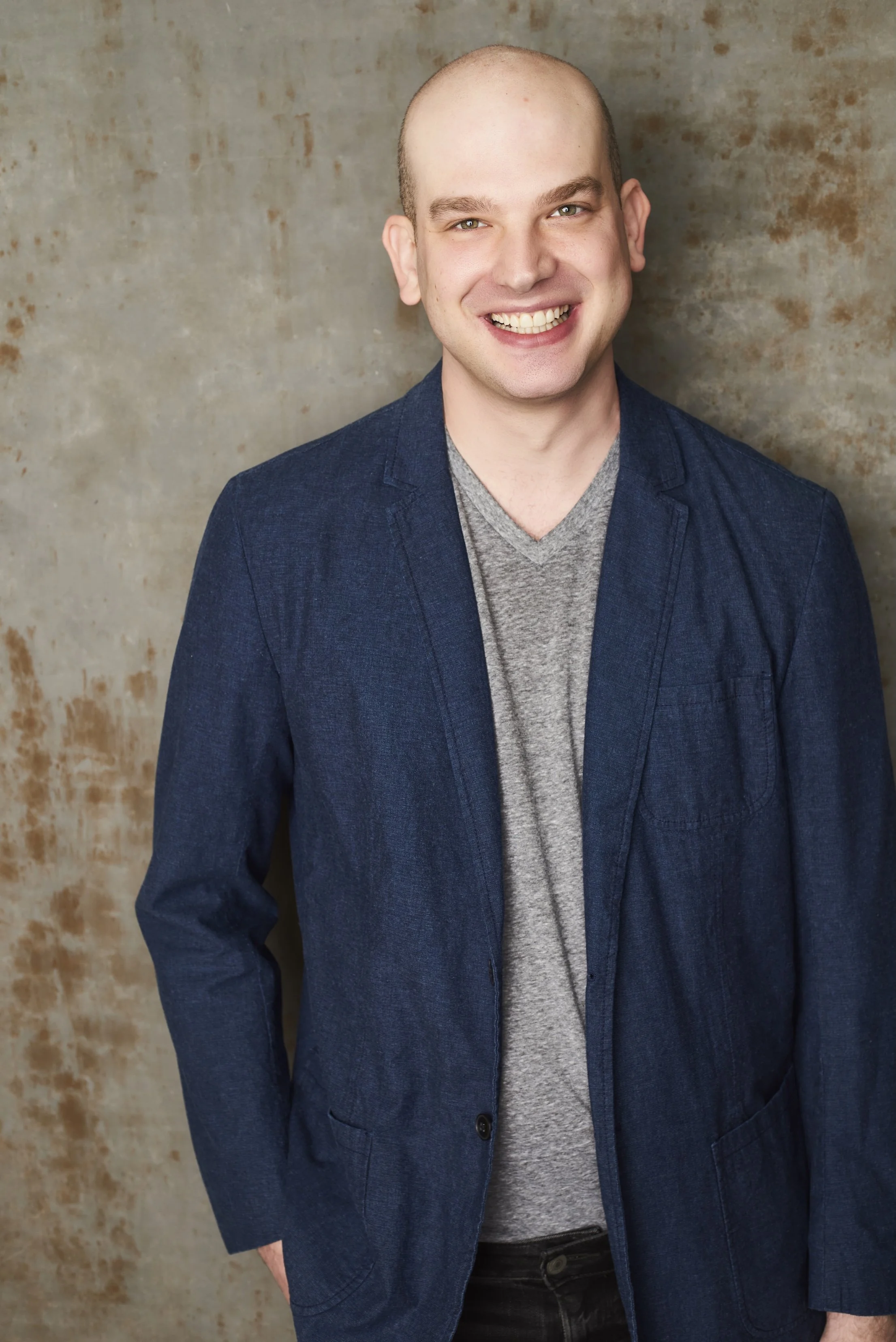 Portrait of a smiling young man with a shaved head wearing a gray t-shirt and navy blazer, standing against a neutral textured background.