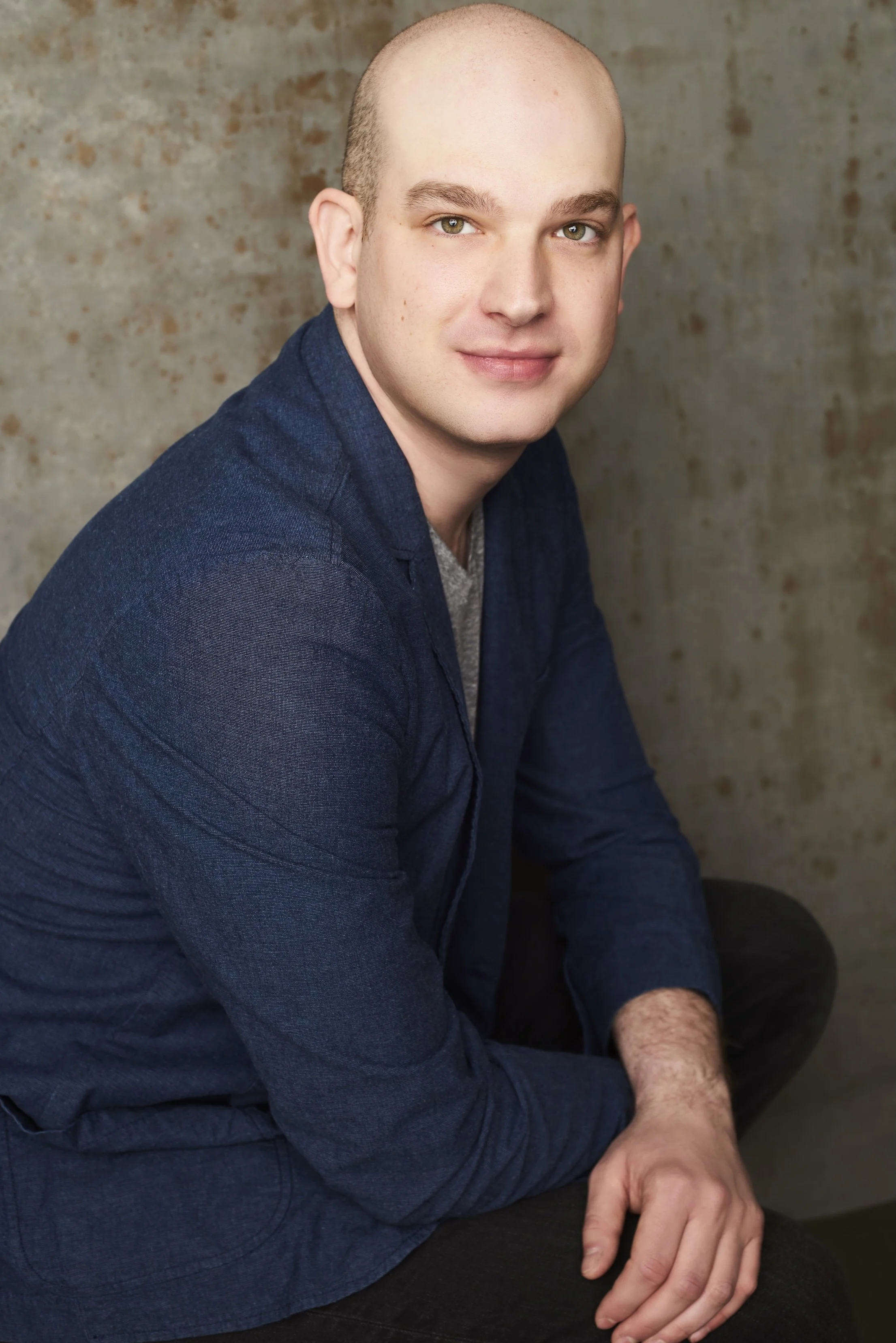 A young man with a bald head and light skin, wearing a navy blue blazer and gray shirt, sitting against a textured beige wall.