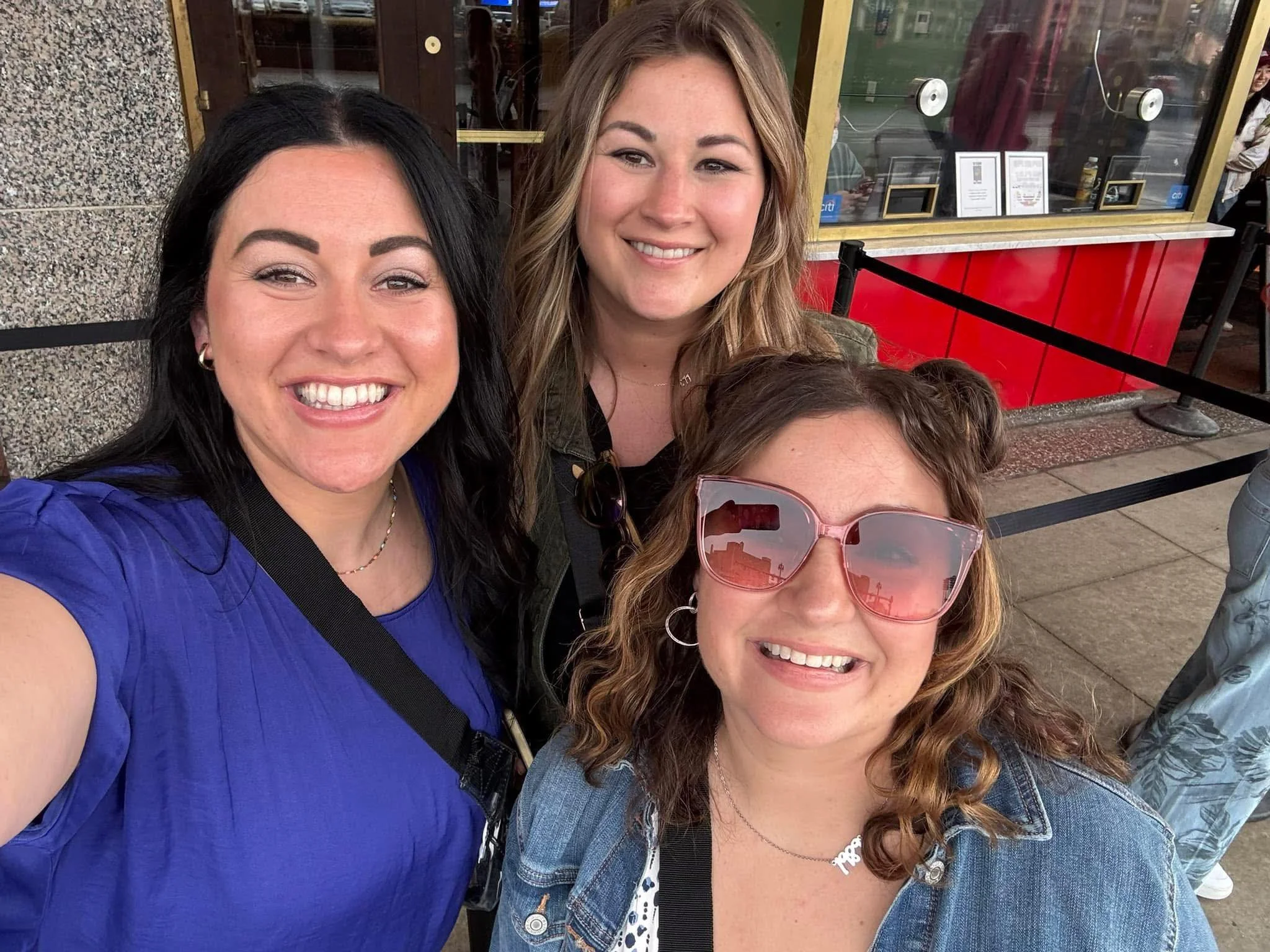Three smiling women taking a selfie outdoors, standing in front of a storefront window with framed photos and signs, on a sidewalk.